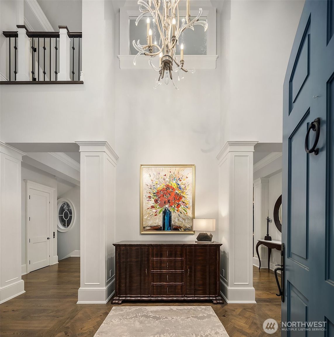 This grand, double-height entryway makes a striking first impression with its soaring ceilings, elegant chandelier, and rich hardwood flooring arranged in a herringbone pattern. A dark wood console table sits centered against the back wall, topped with a vibrant floral painting and a modern lamp, framed by classic white columns. The perspective is from the threshold of the front door, looking into the bright, airy foyer that connects to the rest of the home.