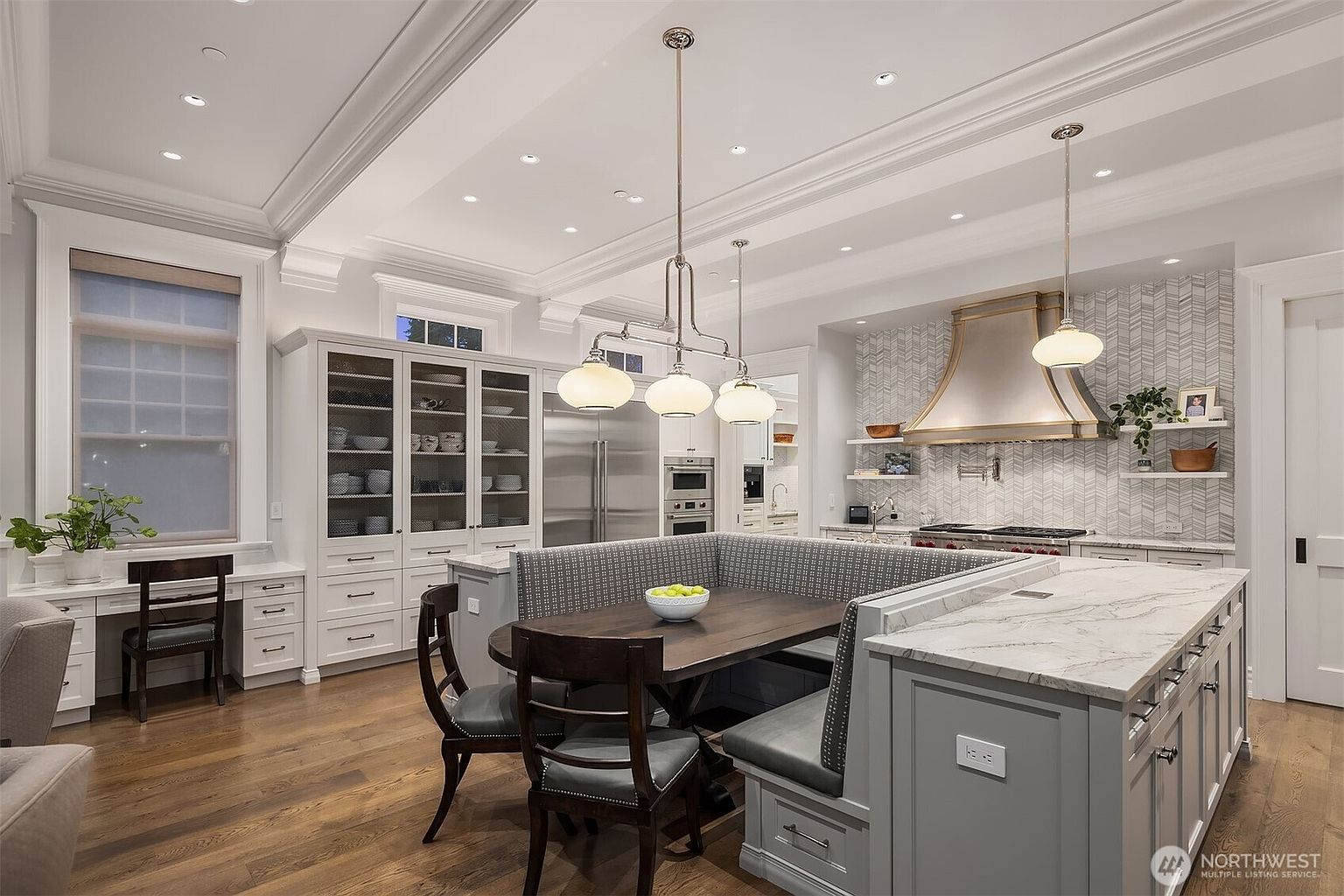 This sophisticated kitchen features a large central island with integrated banquette seating, perfect for casual dining. The space is highlighted by high-end stainless steel appliances, a custom metallic range hood, and elegant herringbone tile backsplash. With its combination of classic cabinetry, marble countertops, and a dedicated built-in desk area, the room offers a seamless blend of luxury and functional design.