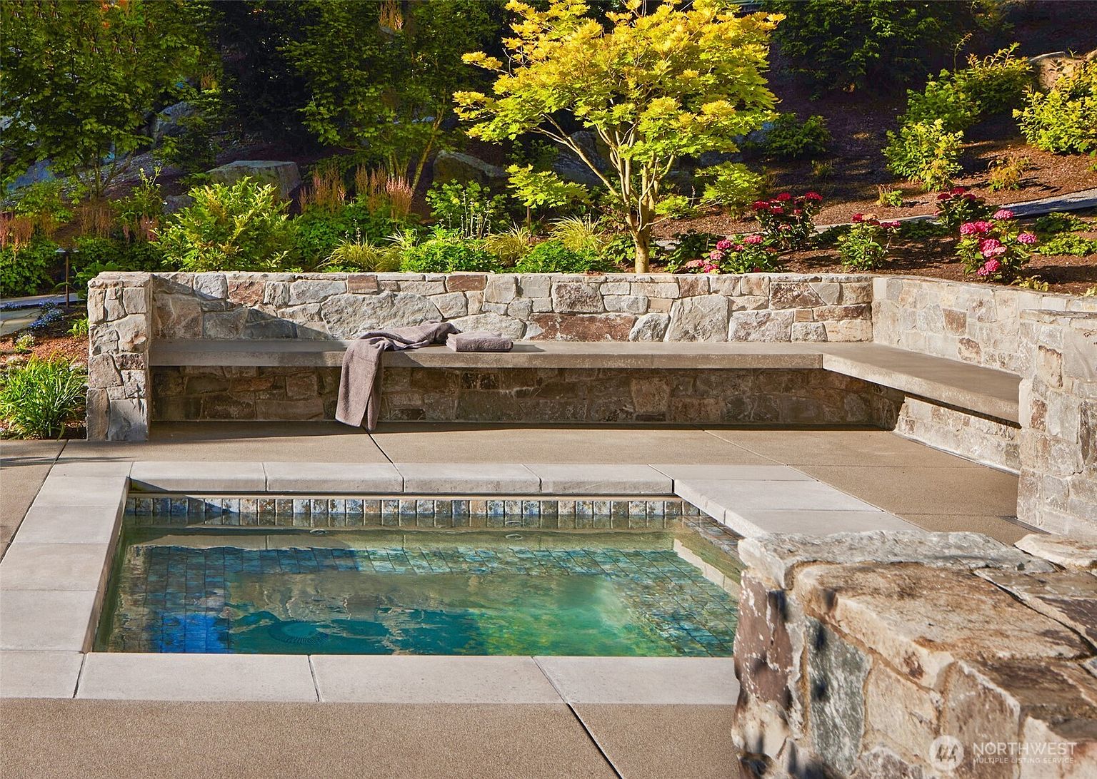 This image showcases a luxurious, custom-built in-ground spa featuring a beautiful mosaic-tiled interior and a smooth concrete coping. The spa is framed by a rustic stone retaining wall that doubles as a built-in bench, creating a sophisticated and relaxing outdoor lounge area. The lush, landscaped garden in the background adds a serene, natural backdrop to this high-end backyard retreat.