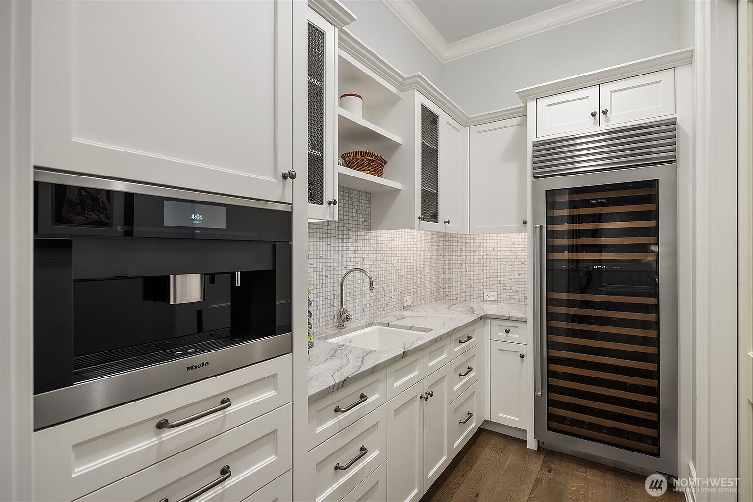This elegant butler's pantry or wet bar area features crisp white cabinetry, marble countertops, and a sophisticated mosaic tile backsplash. The space is equipped with high-end appliances, including a built-in Miele coffee system and a tall Sub-Zero wine refrigerator, creating a luxurious and functional transition space. The perspective is a straight-on, eye-level shot that highlights the clean lines, premium finishes, and organized layout of this high-end home feature.