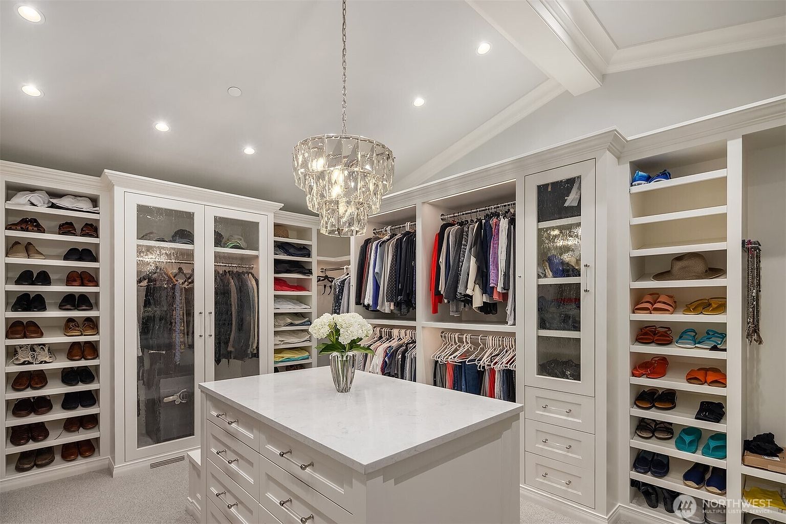 This luxurious walk-in closet features custom white cabinetry, extensive hanging space, and dedicated shelving for shoes and accessories. A central island with a marble countertop and a crystal chandelier serves as a sophisticated focal point, creating an elegant and organized boutique-like atmosphere. The space is well-lit with recessed lighting and carpeted flooring, offering a high-end, functional storage solution.