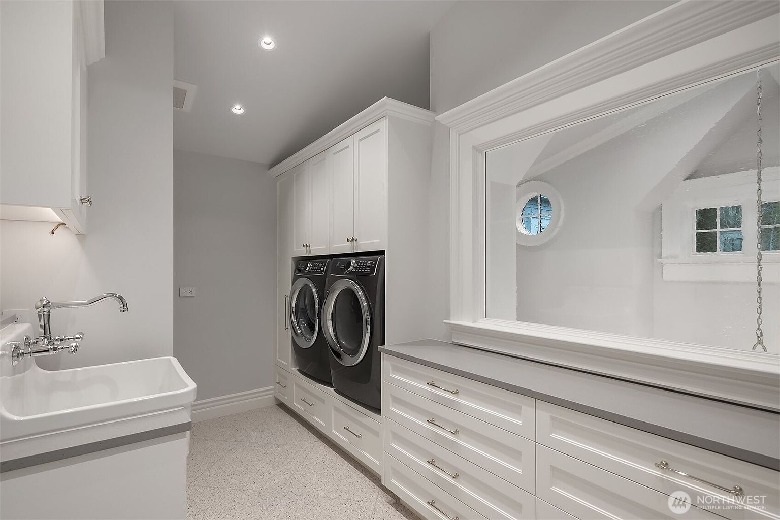 This bright and clean laundry room features a classic aesthetic with white cabinetry, a deep utility sink, and dark-toned front-loading washer and dryer units. A large, framed mirror reflects light across the room, while the speckled flooring adds a subtle texture to the space. The room is well-lit with recessed lighting, creating a functional and polished environment.