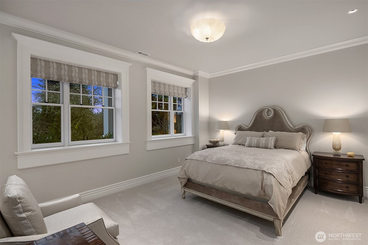 This serene bedroom features a neutral color palette with soft gray walls, plush carpeting, and a sophisticated upholstered headboard. Two large windows with patterned Roman shades allow natural light to fill the space, while matching bedside tables and lamps create a balanced, symmetrical look. The room exudes a calm, elegant atmosphere suitable for a guest suite or secondary bedroom.