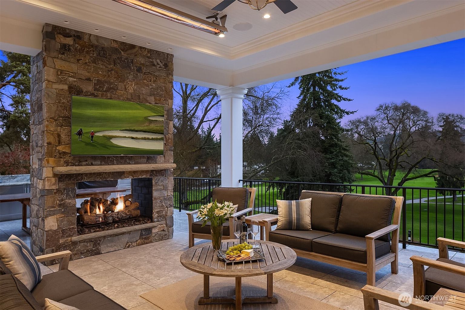 This luxurious covered patio offers a sophisticated outdoor living space, featuring a grand stone fireplace with an integrated television and comfortable lounge seating. The area is furnished with a wooden coffee table holding refreshments and overlooks a lush, manicured golf course at dusk. The elegant design, combined with the serene view, creates an inviting atmosphere perfect for relaxation and entertaining.