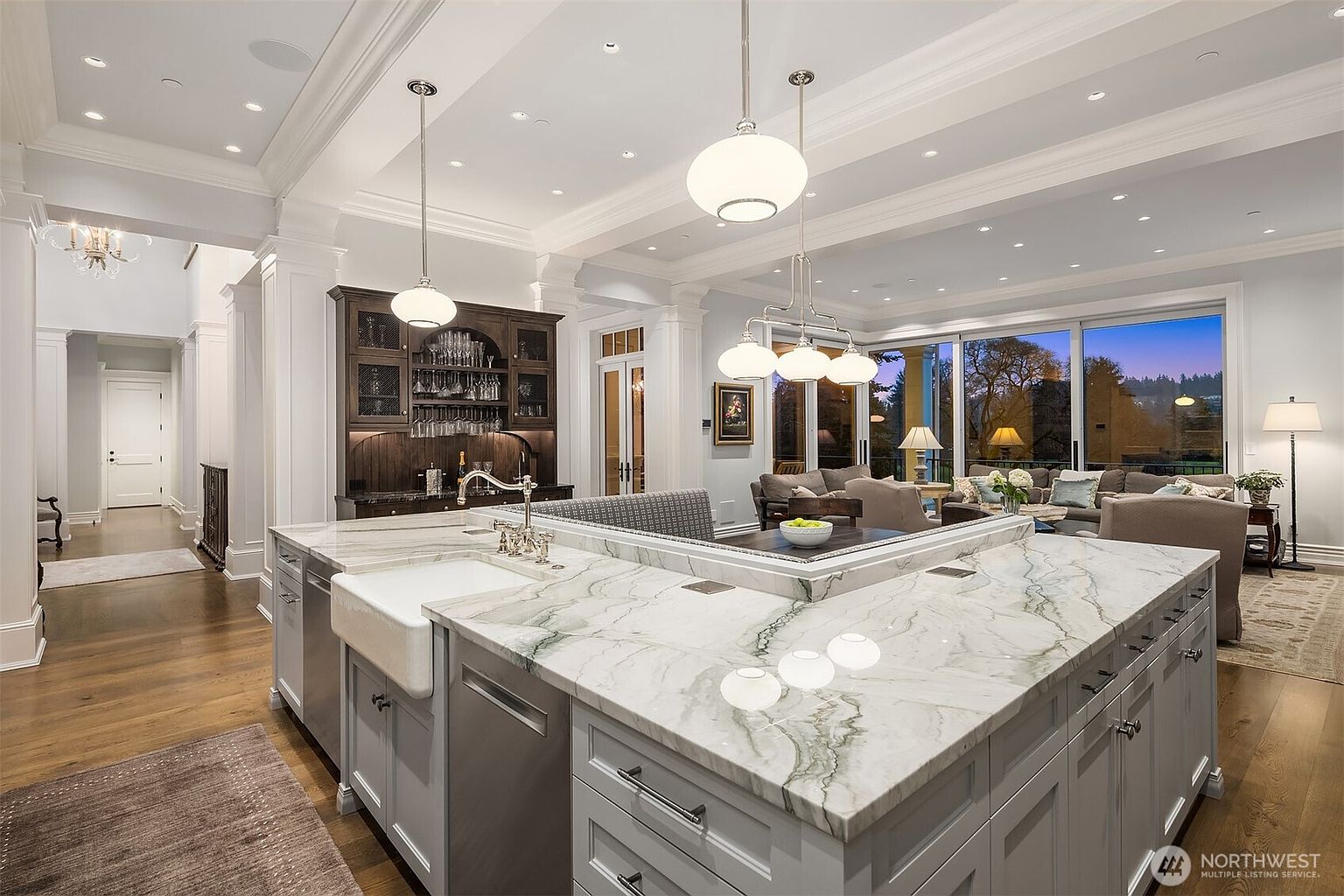 This elegant, open-concept kitchen features a large, marble-topped island with a farmhouse sink and integrated seating, serving as the centerpiece of the room. In the background, a sophisticated wet bar with dark cabinetry complements the bright, airy living area visible through expansive floor-to-ceiling windows. The space is defined by high ceilings, recessed lighting, and a seamless transition between culinary and relaxation zones, creating a luxurious and inviting atmosphere.