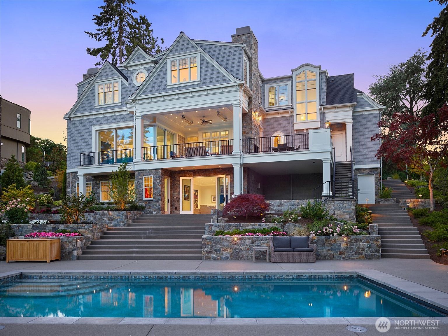 This stunning multi-story luxury residence features a sophisticated blend of stone and shingle siding, showcasing a grand rear elevation that opens onto a tiered stone patio and a pristine swimming pool. The home boasts an expansive covered balcony with ceiling fans, large glass sliding doors, and intricate architectural details, creating an inviting atmosphere for outdoor entertaining. The meticulously landscaped grounds, complete with stone retaining walls and stairs, enhance the property's elegant and high-end aesthetic.