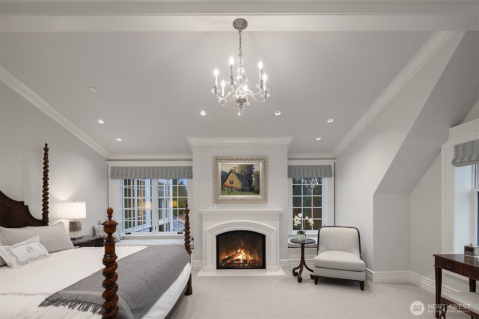 This elegant primary bedroom features a sophisticated neutral color palette, centered around a classic fireplace with a decorative landscape painting above. The room is anchored by a four-poster bed and complemented by a cozy seating area, built-in window benches, and a crystal chandelier that adds a touch of luxury. The space is bright and airy, with large windows that provide natural light and a serene, refined atmosphere.