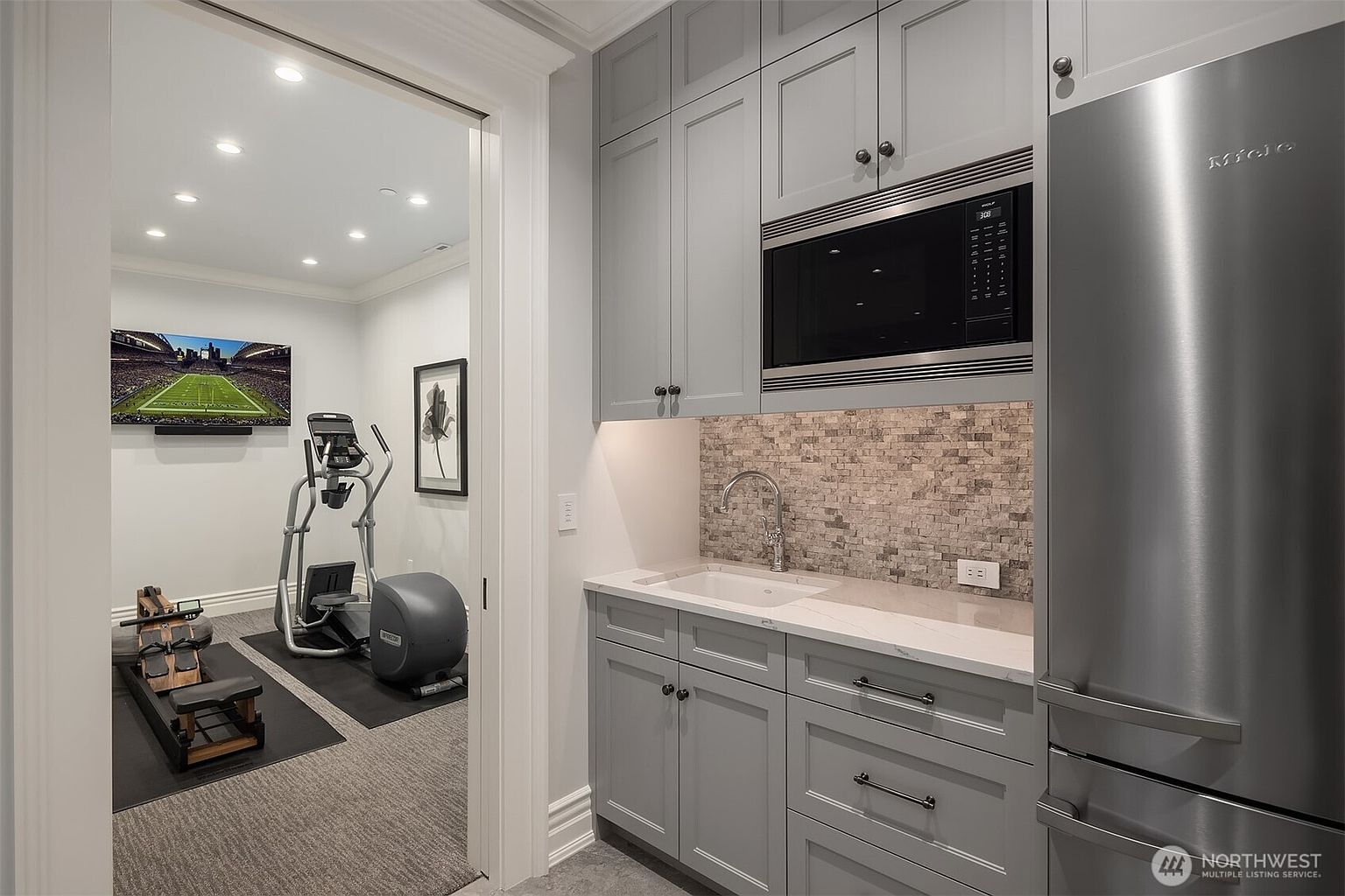 This image captures a multi-functional space featuring a wet bar area in the foreground and a home gym visible through an open doorway. The wet bar is equipped with grey cabinetry, a stone backsplash, a sink, and a built-in microwave, while the adjacent gym contains professional-grade fitness equipment including a rowing machine and an elliptical. The perspective creates a seamless transition between a convenient refreshment station and a dedicated workout area, showcasing a sophisticated and practical home layout.