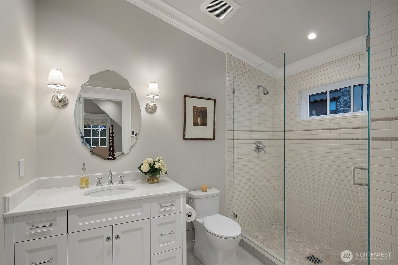 This bright and clean primary bathroom features a white vanity with a marble countertop, a stylish oval mirror, and dual wall-mounted light fixtures. To the right, a spacious walk-in shower is enclosed by a glass panel and finished with classic subway tile and a pebble-stone floor. The neutral color palette and modern fixtures create a serene and inviting atmosphere.