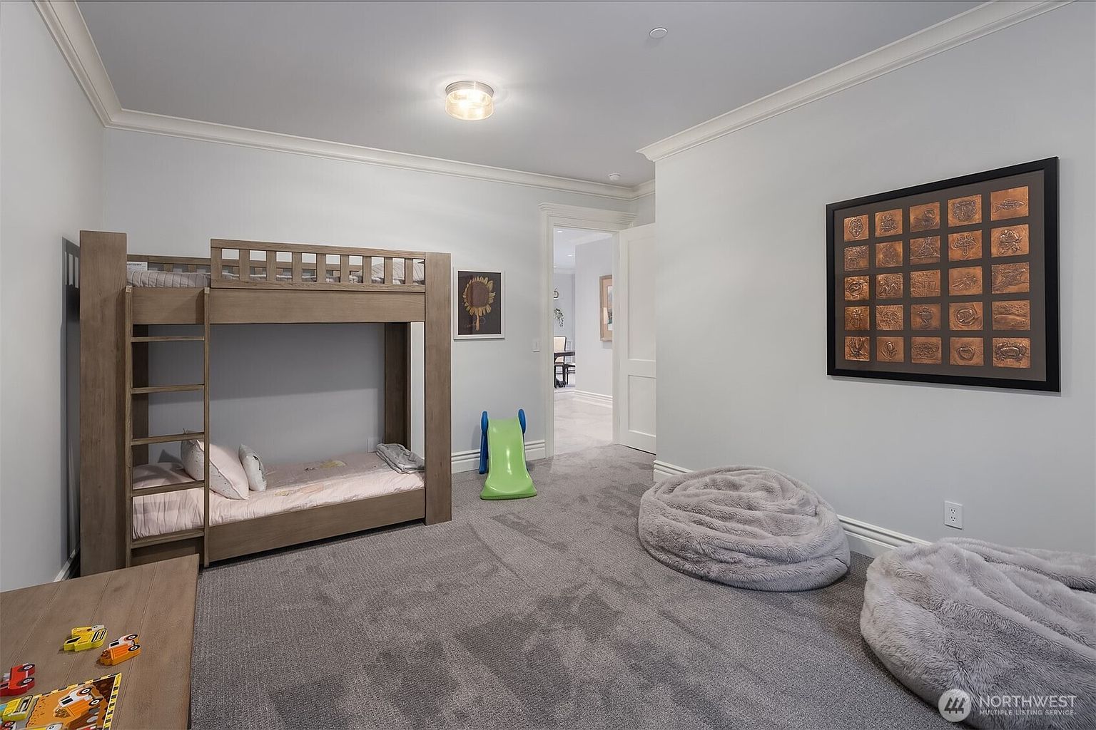 This spacious children's bedroom features a light-toned wooden bunk bed with an integrated slide, creating a playful and functional atmosphere. The room is carpeted in a neutral grey, complemented by two large, plush beanbag chairs and a framed art piece on the wall. The perspective is from the doorway, looking into the room, which leads to an adjacent hallway, highlighting the home's open and inviting layout.