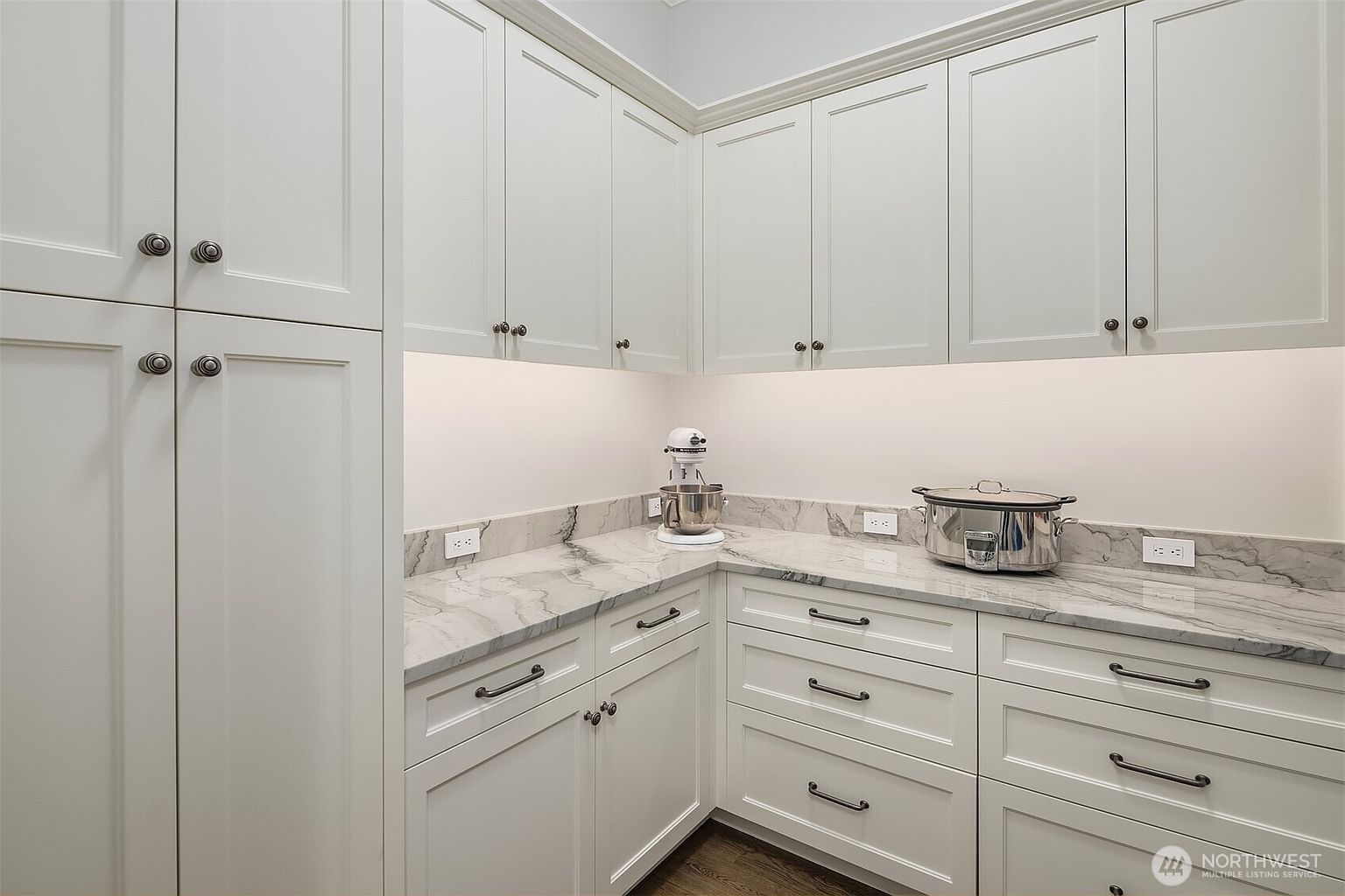This well-appointed butler's pantry or secondary kitchen area features crisp white shaker-style cabinetry paired with elegant marble-patterned countertops. The space is illuminated by under-cabinet lighting, highlighting the clean lines and modern hardware, while a stand mixer and slow cooker sit on the counter, suggesting a functional and organized workspace. The perspective is a straight-on, eye-level shot that emphasizes the efficient layout and high-end finishes of the room.