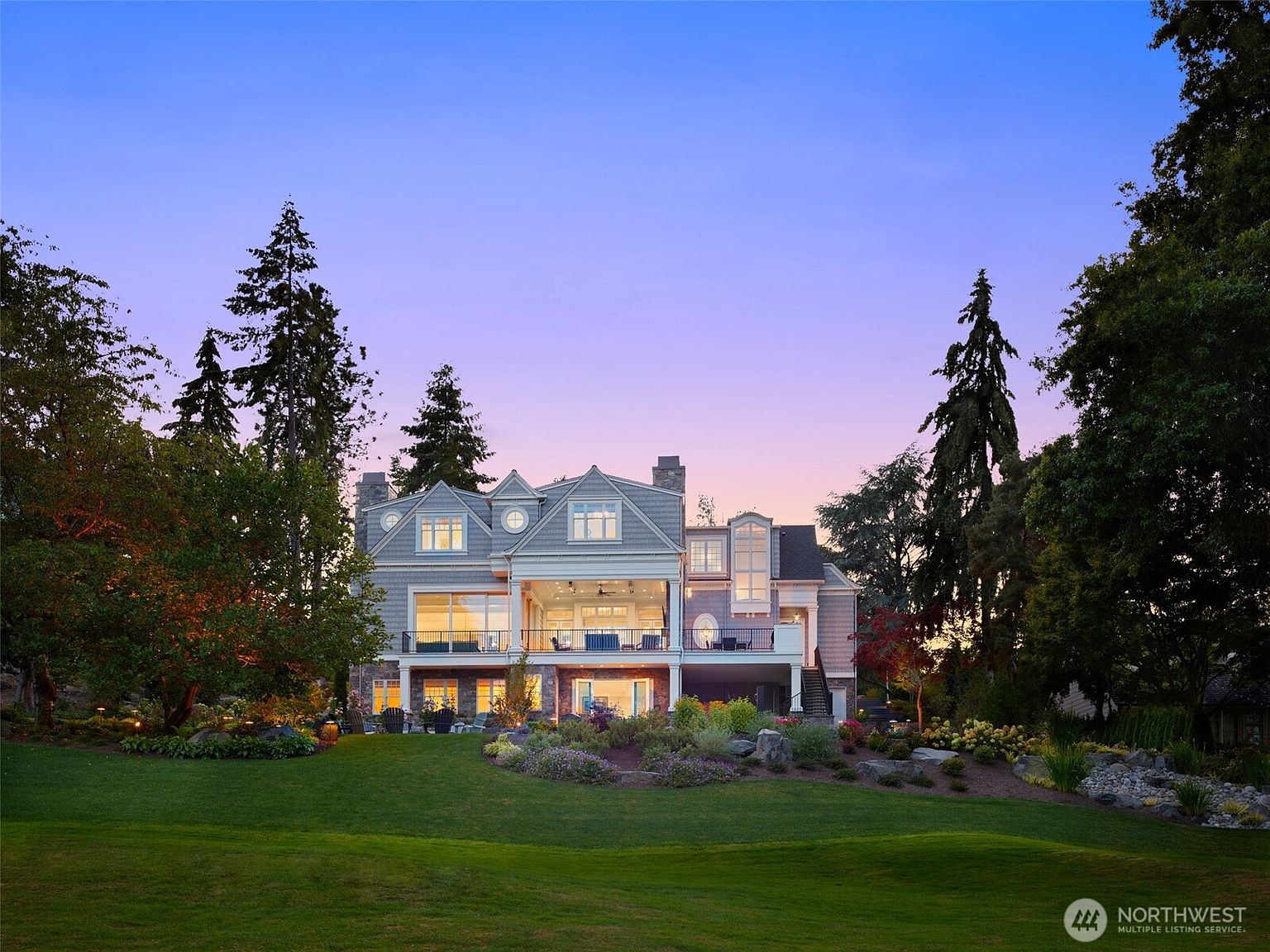 This stunning rear exterior view captures a multi-story luxury residence at dusk, showcasing a blend of shingle-style architecture and stone masonry. The home features a spacious elevated deck with integrated lighting, large glass sliding doors, and a beautifully landscaped backyard with lush green grass and rock garden accents. The twilight sky provides a serene backdrop, highlighting the warm, inviting glow emanating from the home's interior.