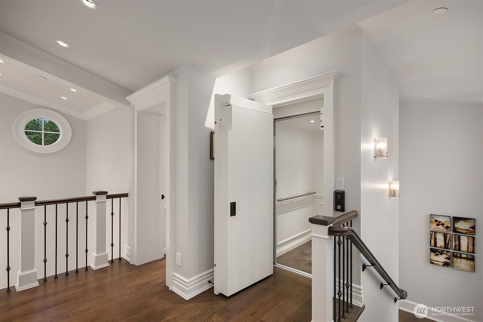 This interior view captures a sophisticated landing area featuring a prominent residential elevator with a white door, seamlessly integrated into the home's architecture. To the left, a classic staircase with dark wood railings leads downward, while a unique circular window adds a touch of architectural character to the space. The neutral color palette and polished hardwood floors create an elegant, high-end atmosphere suitable for a luxury residence.