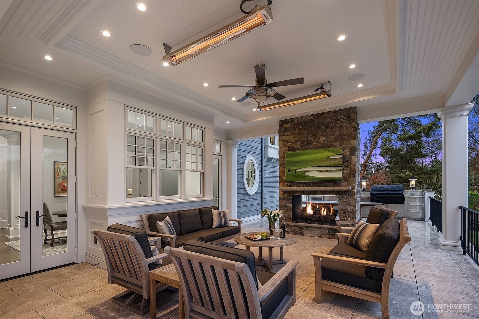 This luxurious covered outdoor patio features a stone fireplace with an integrated television, creating a cozy and sophisticated living space. The area is furnished with comfortable dark-cushioned seating, a wooden coffee table, and overhead heaters, all set against a backdrop of elegant white trim and large windows. The perspective captures a welcoming, high-end outdoor retreat perfect for entertaining, with a glimpse of the surrounding landscape at dusk.