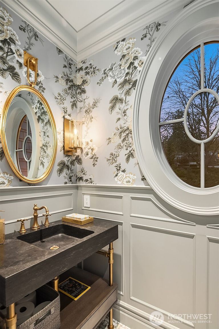 This elegant powder room features sophisticated floral wallpaper in soft grey and white tones, complemented by classic wainscoting. A striking dark stone vanity with brass plumbing fixtures and a matching oval mirror creates a luxurious focal point, while a large, circular window allows natural light to flood the space. The overall aesthetic is refined, blending traditional architectural details with modern, high-end finishes.