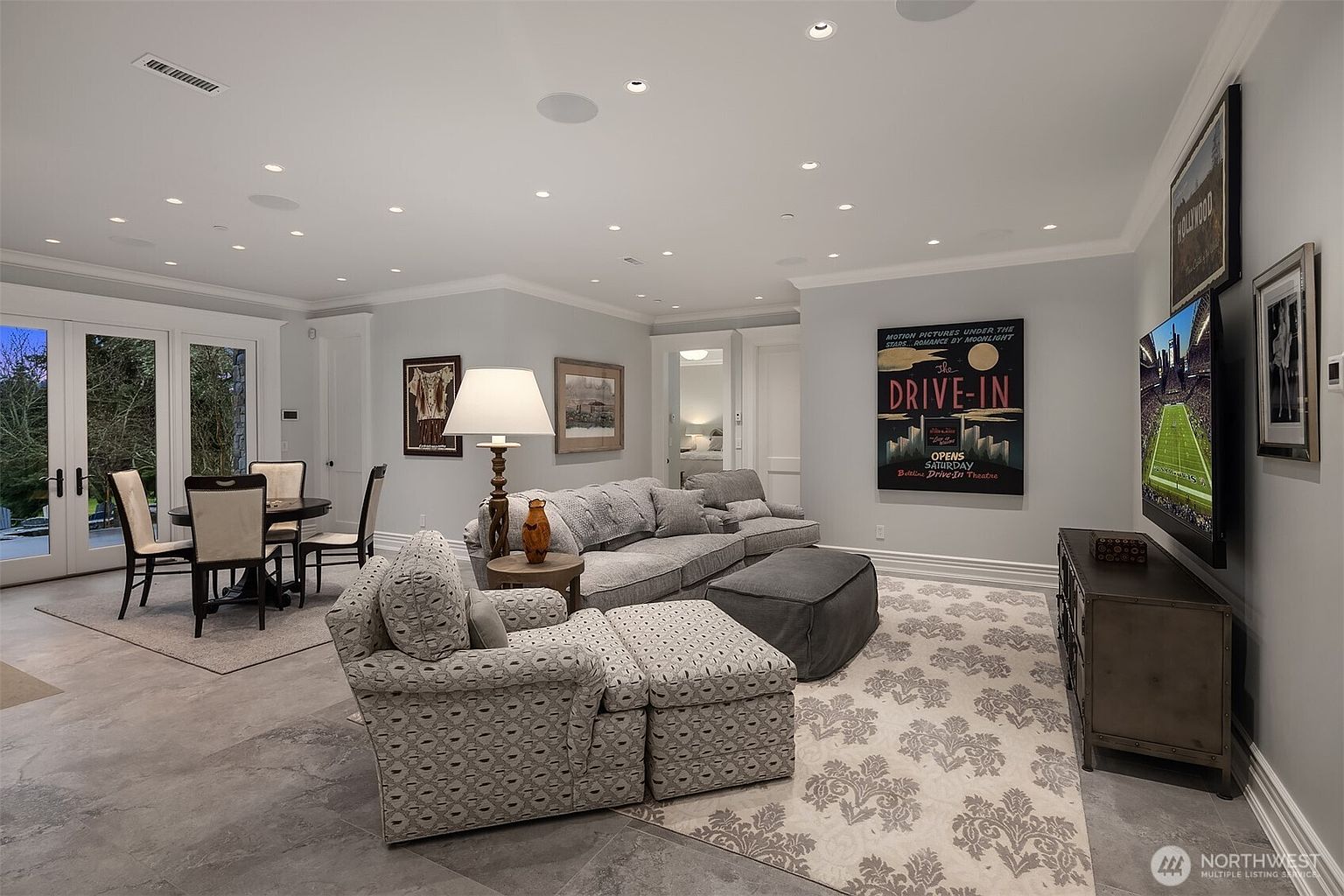 This spacious, open-concept living room features a comfortable gray sectional sofa, a matching patterned armchair with an ottoman, and a large area rug over tiled flooring. The room is decorated with framed artwork, including a vintage-style 'Drive-In' poster, and includes a dining area near the glass patio doors. Recessed lighting and a large wall-mounted television create a cozy yet sophisticated atmosphere perfect for relaxation and entertainment.