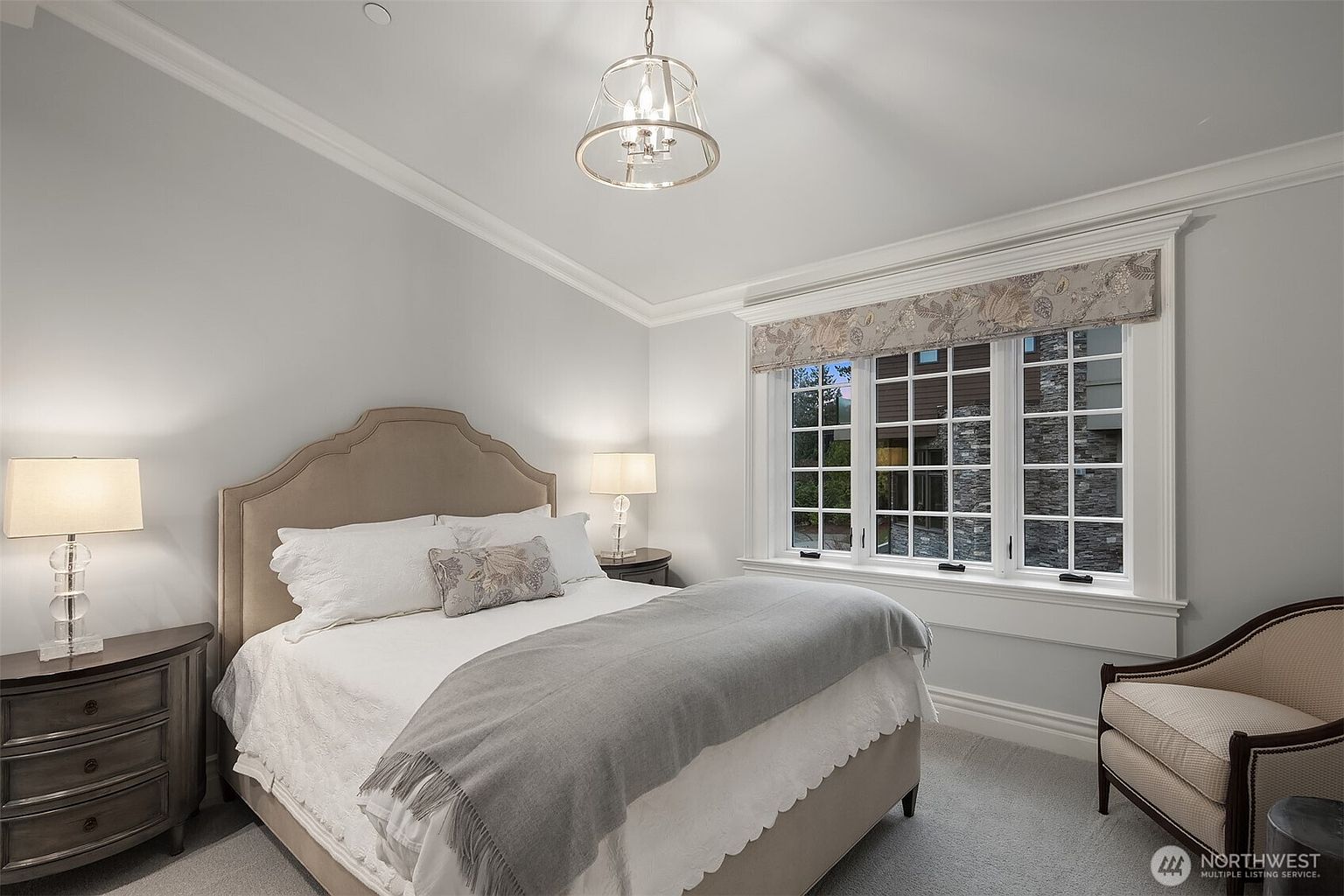 This elegant bedroom features a neutral color palette with a plush upholstered headboard and a cozy grey throw blanket draped over the bed. A large window with a patterned valance allows natural light to fill the room, while a sophisticated chandelier provides a touch of luxury. The space is completed by matching bedside tables with glass lamps and a comfortable armchair, creating a serene and inviting atmosphere.
