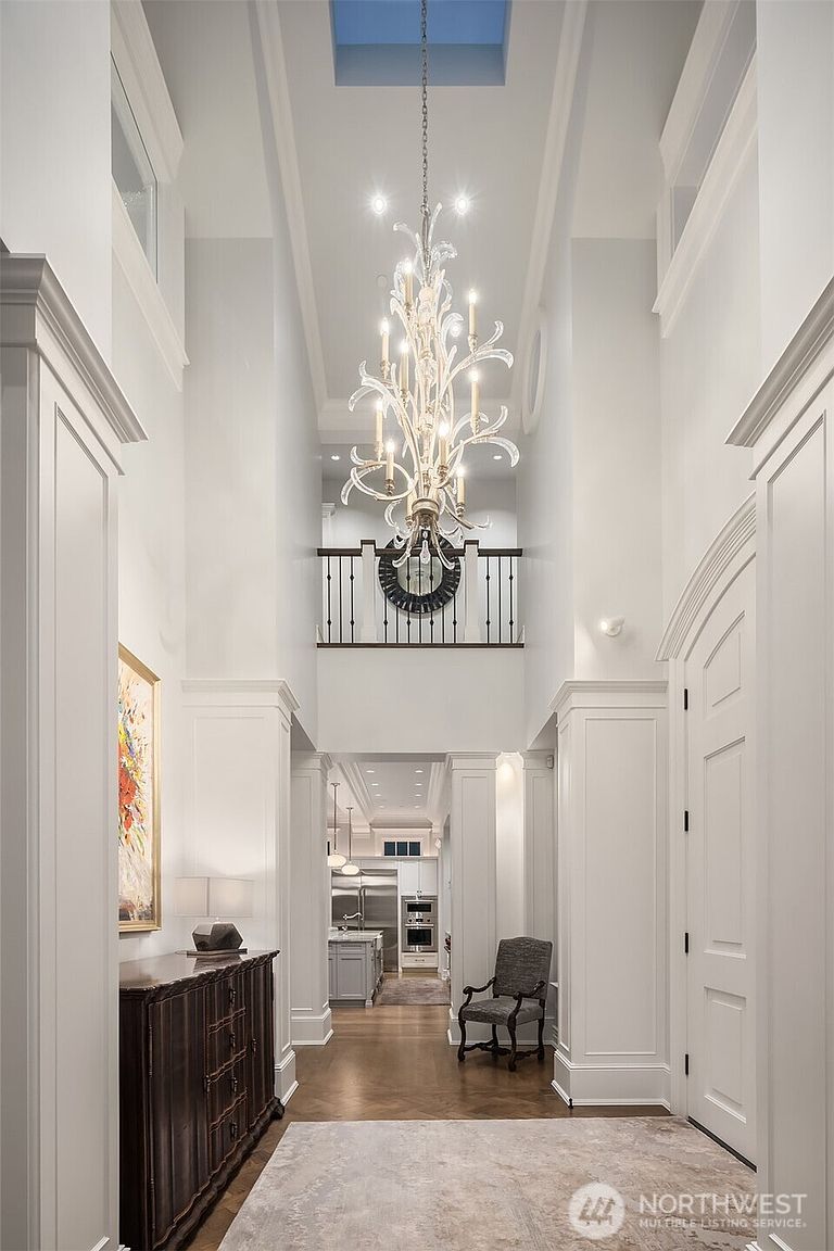 This grand, double-height entryway features a dramatic crystal chandelier suspended from a high ceiling with a skylight, creating an airy and luxurious atmosphere. The space is defined by crisp white wainscoting, dark hardwood floors, and a sophisticated landing with a decorative iron railing overlooking the foyer. A dark wood console table and a classic armchair add a touch of traditional elegance, while the perspective draws the eye through the hallway toward the bright, modern kitchen in the background.