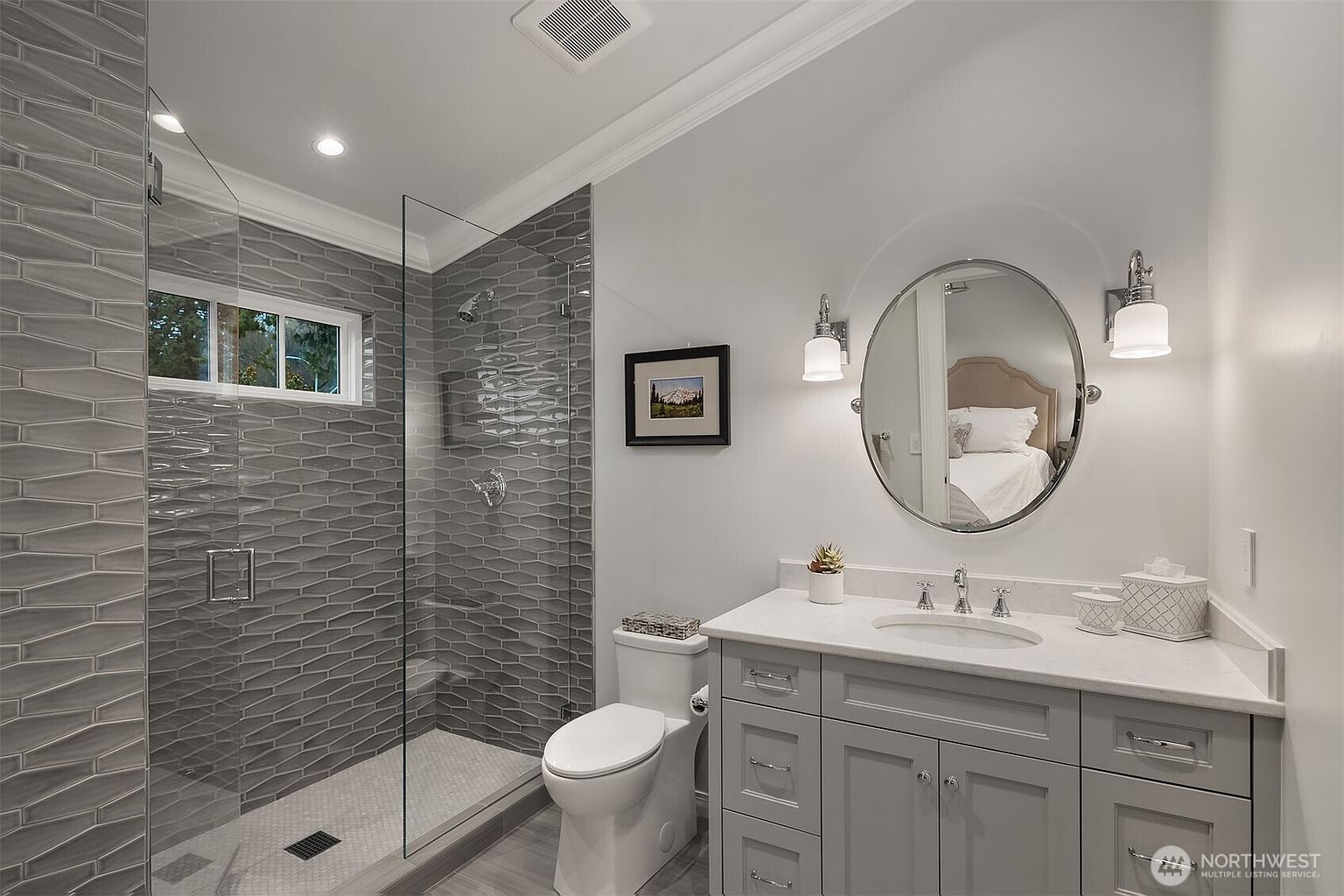 This modern bathroom features a sleek gray vanity with a white countertop, a circular mirror, and elegant wall-mounted light fixtures. To the left, a spacious walk-in shower is enclosed by a glass panel and finished with textured, elongated gray tiles. The room is bright and clean, offering a contemporary aesthetic that feels both functional and stylish.