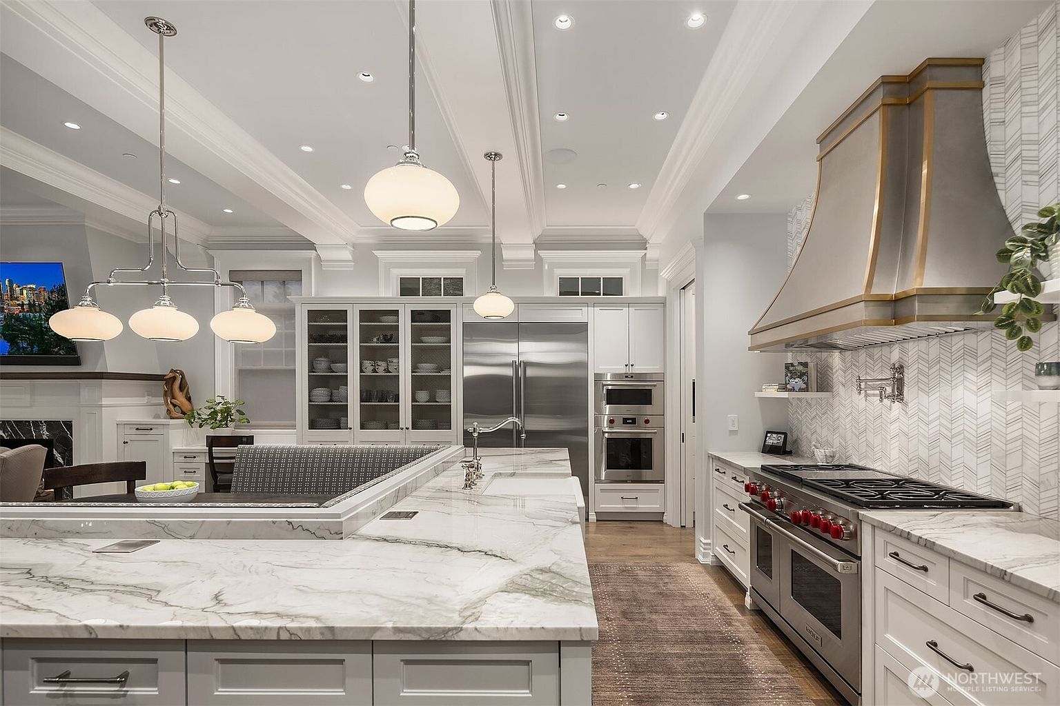 This sophisticated, high-end kitchen features a massive marble-topped island with integrated seating, serving as the centerpiece of the open-concept space. The room is anchored by a professional-grade Wolf range with a custom metallic hood, complemented by sleek white cabinetry, stainless steel appliances, and elegant pendant lighting. The perspective captures the seamless flow between the culinary workspace and the adjacent living area, highlighting a luxurious and functional design.