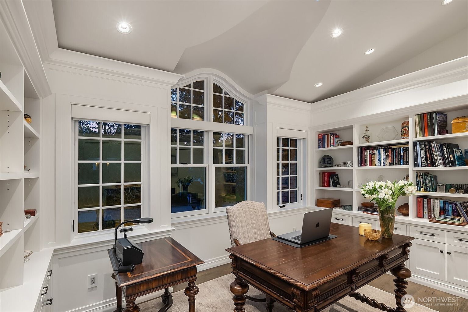 This elegant home office features custom built-in white bookshelves, a rich dark wood desk, and a sophisticated, traditional aesthetic. Large, multi-paned windows with an arched top allow for abundant natural light, creating a bright and productive atmosphere. The room is accented by a comfortable upholstered chair, a laptop, and a vase of fresh flowers, making it a refined and inviting workspace.