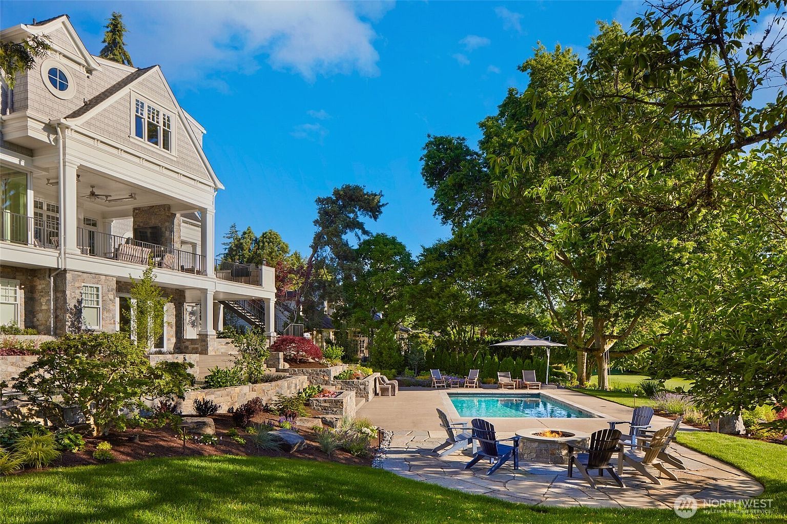 This stunning rear exterior view showcases a luxurious multi-story home with a spacious covered patio and a beautifully landscaped backyard. The scene features a pristine rectangular swimming pool, a stone fire pit area with Adirondack chairs, and lush greenery, creating an inviting and serene outdoor living space. The perspective captures the seamless transition between the elegant architecture of the house and the expansive, well-manicured lawn.