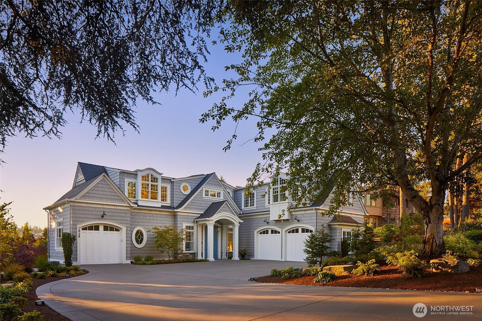 This stunning, custom-built home features classic shingle-style architecture with a sophisticated grey exterior and crisp white trim. The property boasts a spacious, curved concrete driveway leading to a three-car garage, complemented by mature trees and manicured landscaping that frame the elegant entryway. The warm, golden-hour lighting enhances the inviting curb appeal, highlighting the home's symmetrical design and architectural charm.