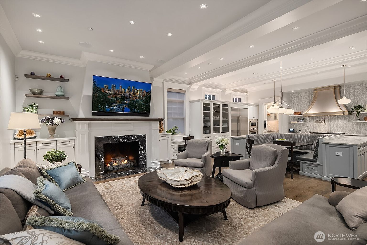 This elegant open-concept living room features a sophisticated fireplace with a black marble surround and a large wall-mounted television. The space is furnished with plush grey seating, a dark wood oval coffee table, and built-in shelving, seamlessly transitioning into a cozy kitchen dining nook. The room is bathed in warm, recessed lighting, creating an inviting and luxurious atmosphere perfect for entertaining.