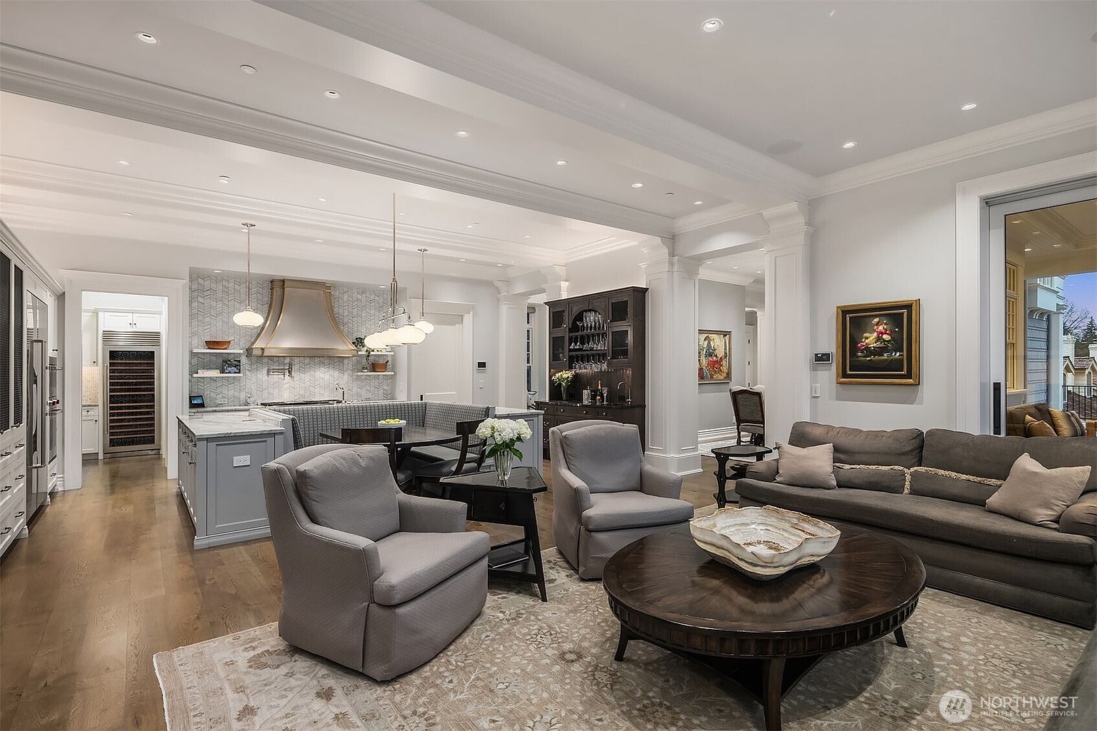 This elegant open-concept living space seamlessly blends a cozy lounge area with a sophisticated kitchen and dining nook. The room features high ceilings with recessed lighting, rich hardwood floors, and a neutral color palette that highlights the custom cabinetry and high-end finishes. A large, dark wood coffee table serves as the centerpiece, while the adjacent kitchen area boasts a stylish herringbone backsplash and a functional breakfast banquette.