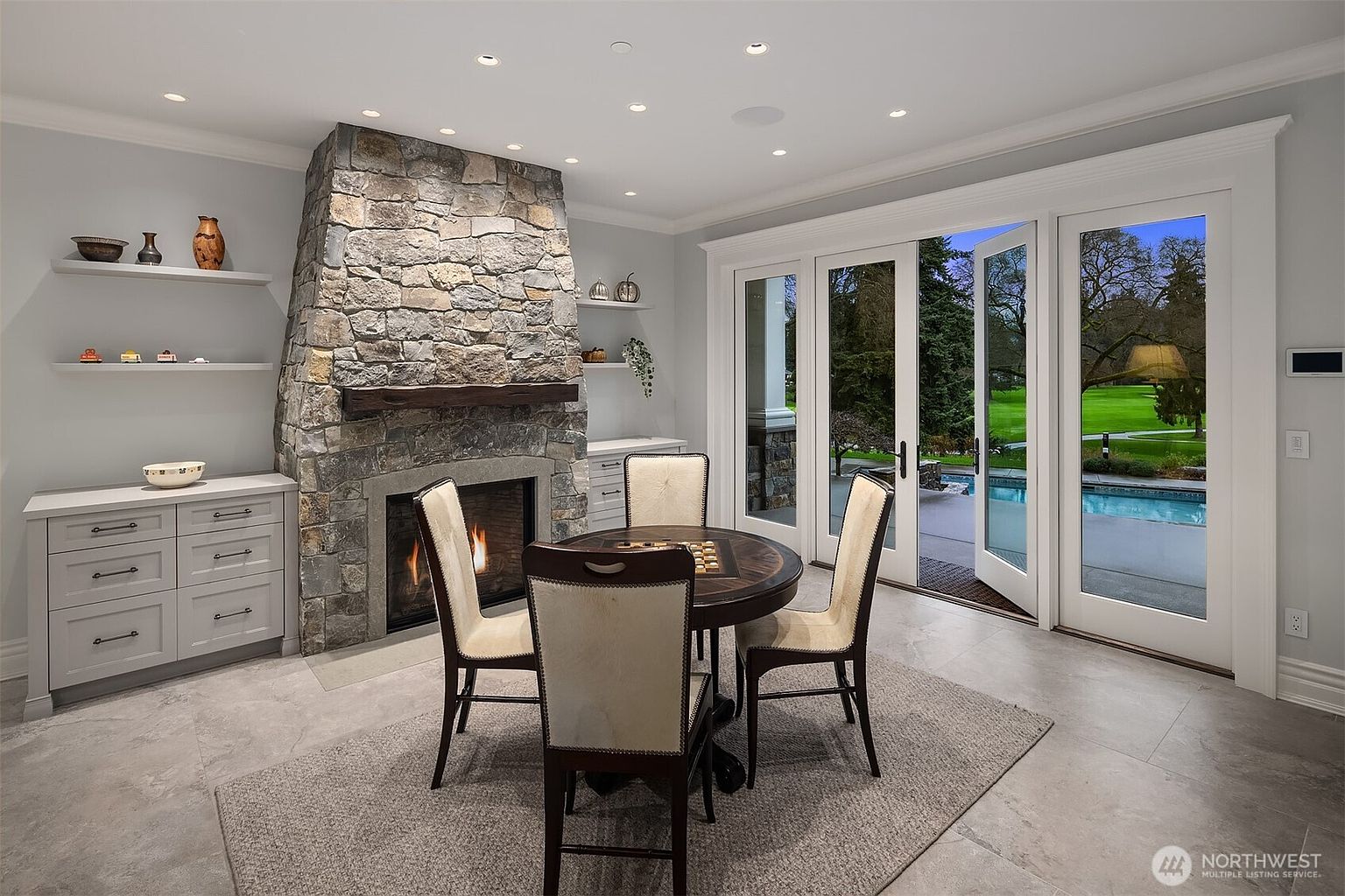 This elegant dining area features a prominent stone fireplace with a rustic wooden mantel, serving as the room's focal point. A circular table with four upholstered chairs sits atop a textured area rug, while French doors open to a scenic view of a backyard pool and lush greenery. The space is finished with light grey cabinetry and built-in shelving, creating a sophisticated and inviting atmosphere.