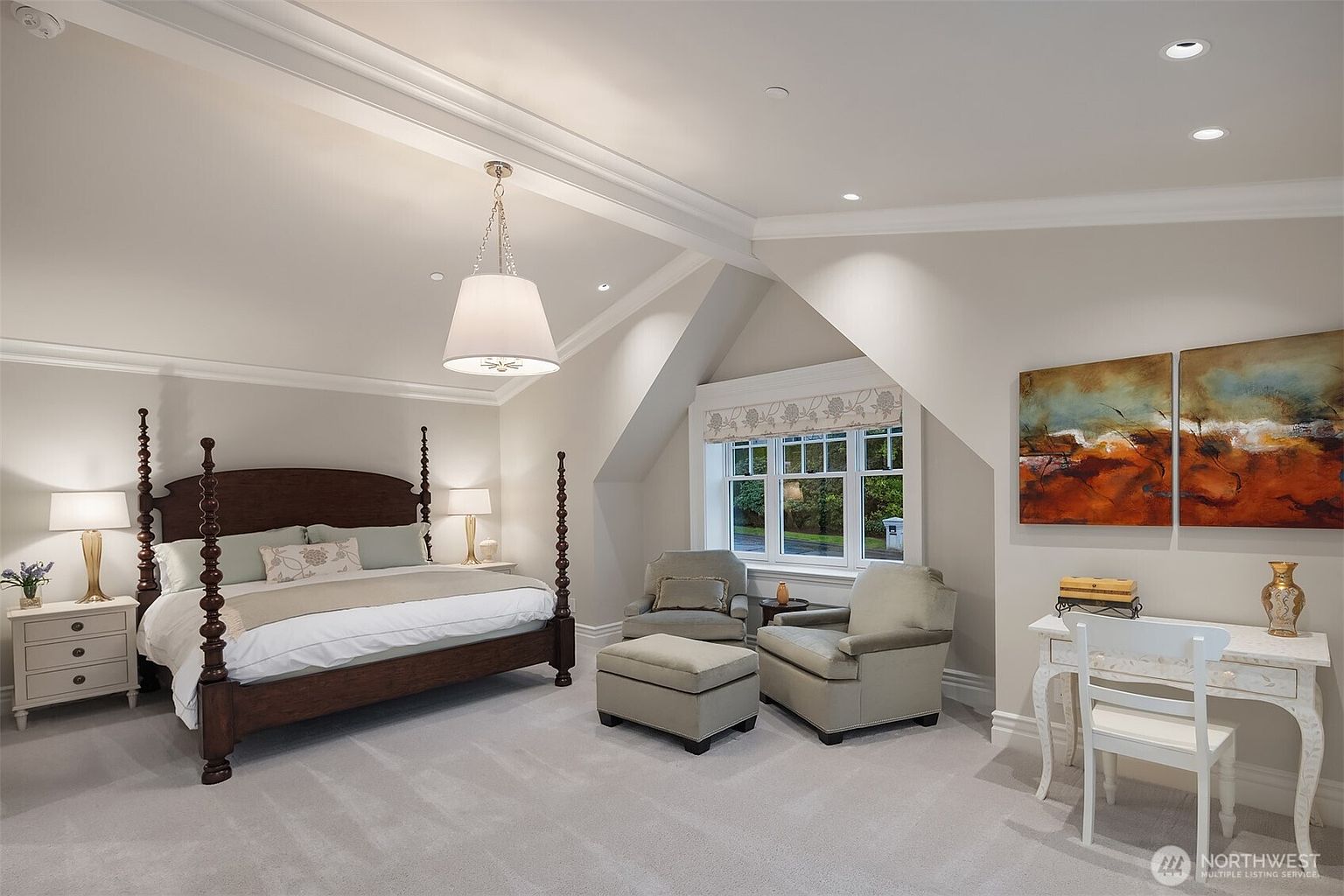 This spacious primary bedroom features a grand four-poster wooden bed as the focal point, complemented by a cozy seating area with two armchairs and an ottoman. The room is bathed in natural light from a large window, while elegant crown molding and a sophisticated chandelier add a touch of luxury. A dedicated desk area and tasteful wall art complete the serene and well-appointed atmosphere.