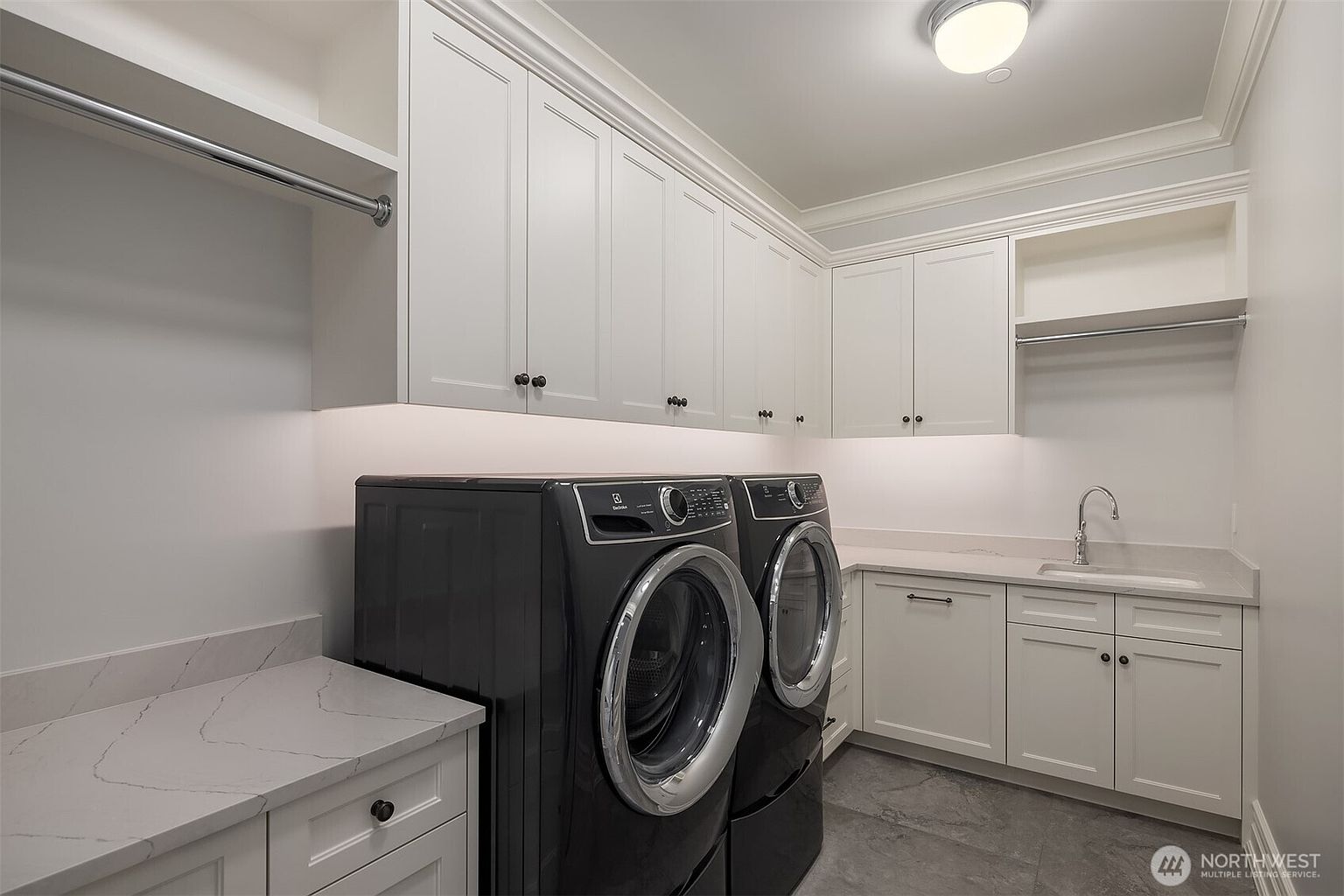 This modern, well-organized laundry room features sleek white cabinetry, quartz countertops, and a pair of high-end black front-load appliances. The space includes a built-in sink, under-cabinet lighting, and a hanging rod, creating a functional and polished aesthetic. The perspective is a wide-angle shot that captures the efficient layout and clean, contemporary design of the utility area.