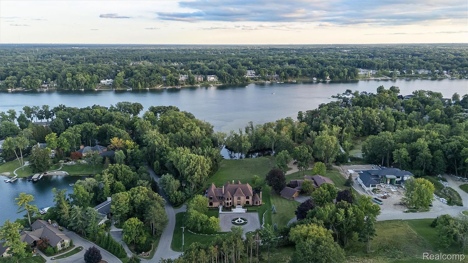 This aerial shot showcases a luxurious estate nestled among lush greenery and bordering a serene lake. The property features a grand residence with manicured lawns, a circular driveway, and mature trees, complemented by neighboring upscale homes and waterfront access. The overall impression is one of exclusivity and natural beauty.