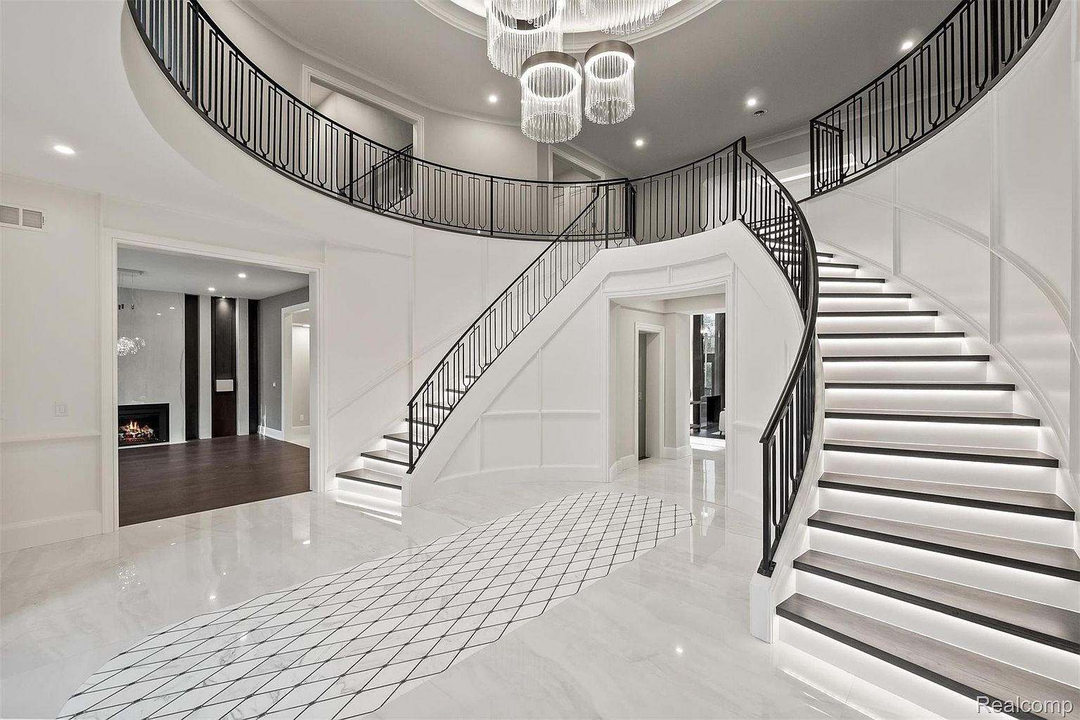 This grand foyer features a sweeping staircase with black metal railings and integrated lighting, creating a dramatic and luxurious entrance. The white marble flooring with a geometric inlay adds to the elegance, while the open layout provides a sense of spaciousness. A modern chandelier hangs from the ceiling, enhancing the sophisticated ambiance.