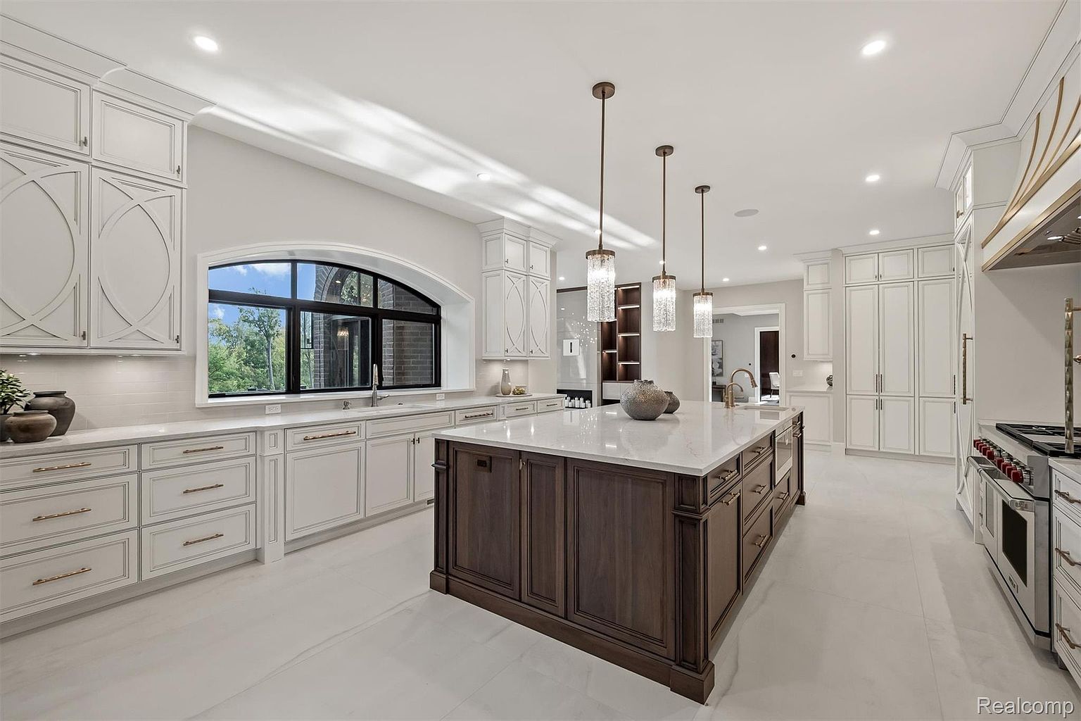 This is a bright and spacious kitchen featuring custom white cabinetry, a large dark wood island with a white countertop, and stainless steel appliances. The kitchen is well-lit with pendant lighting and natural light from a large arched window. The overall design is elegant and modern, creating a luxurious and functional space.