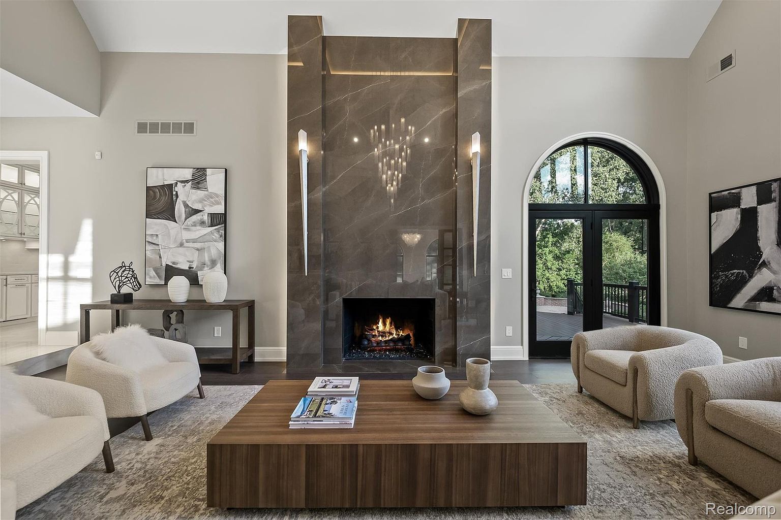 This is an interior shot of a luxurious living room featuring a grand fireplace with marble accents and modern lighting fixtures. The room is furnished with plush armchairs, a large wooden coffee table, and abstract artwork, creating a sophisticated and inviting atmosphere. A large arched doorway leads to an outdoor deck, enhancing the sense of space and connection to nature.