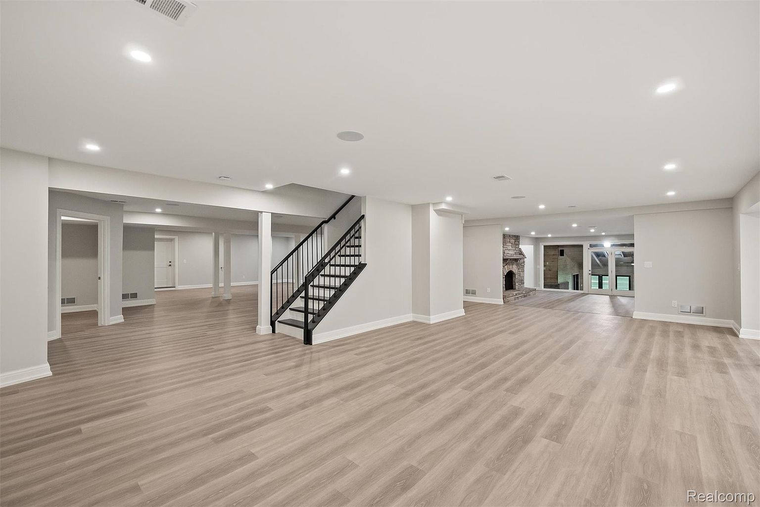 This is a spacious, newly renovated basement featuring light wood-look flooring and neutral gray walls. The open layout includes a staircase with black metal railings, a stone fireplace, and recessed lighting throughout. The space is bright and airy, offering a versatile area for recreation or entertainment.