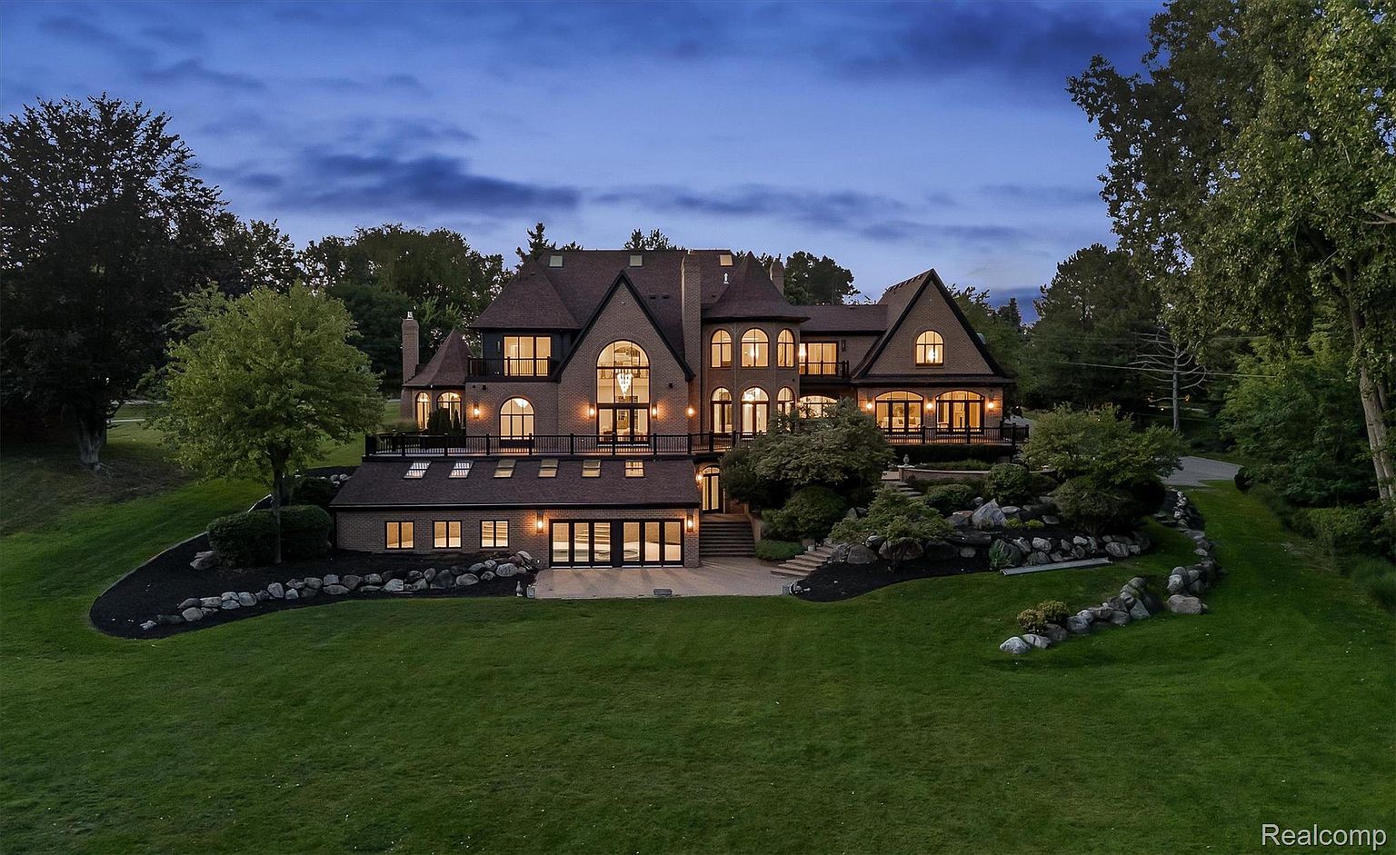 This is a stunning front exterior view of a large, luxurious home at dusk. The house features a brick facade, multiple arched windows, a dark roof, and a well-manicured lawn with rock landscaping. The warm interior lights create an inviting ambiance, highlighting the architectural details and grandeur of the property.