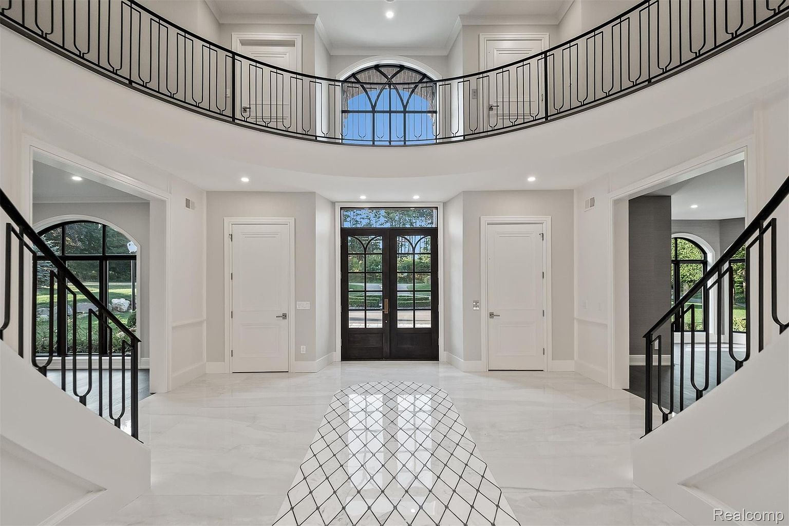 This grand foyer features a soaring two-story design with a curved balcony overlooking the entrance. The space is illuminated by recessed lighting and natural light streaming through the front doors and upper window. A striking black and white tile design leads into the home, flanked by staircases with black railings and white risers.