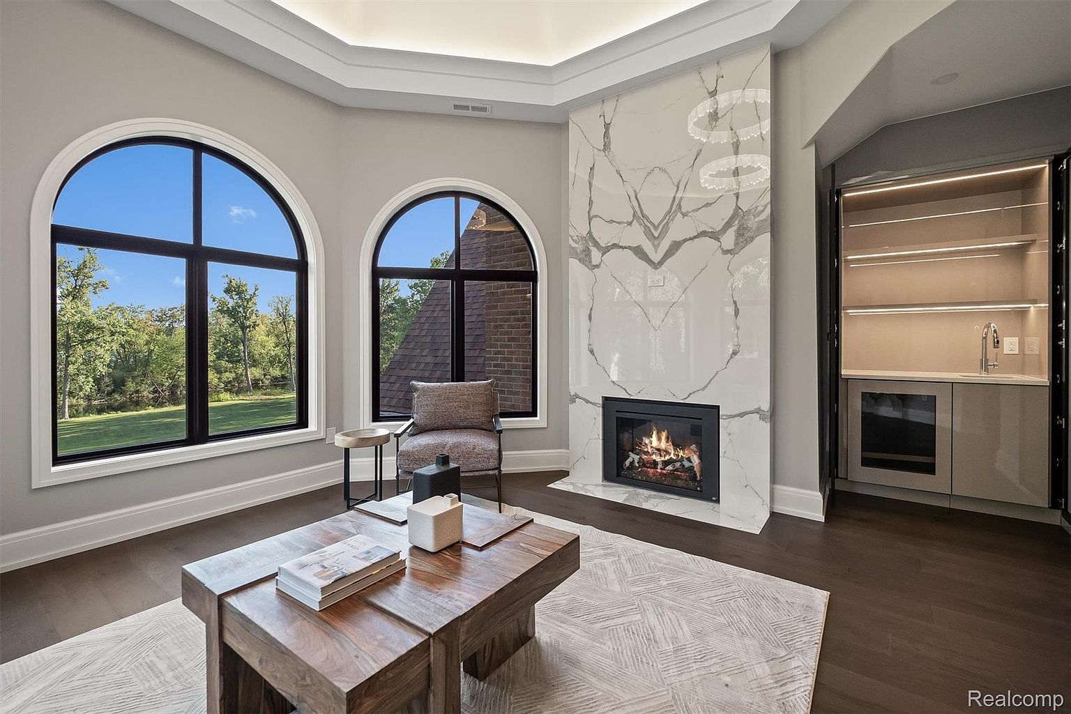 This interior shot showcases a luxurious living room with dark hardwood floors and a striking marble fireplace. Large arched windows provide ample natural light and views of the exterior greenery. The room is furnished with a modern coffee table, a stylish armchair, and a built-in bar area, creating an inviting and sophisticated atmosphere.
