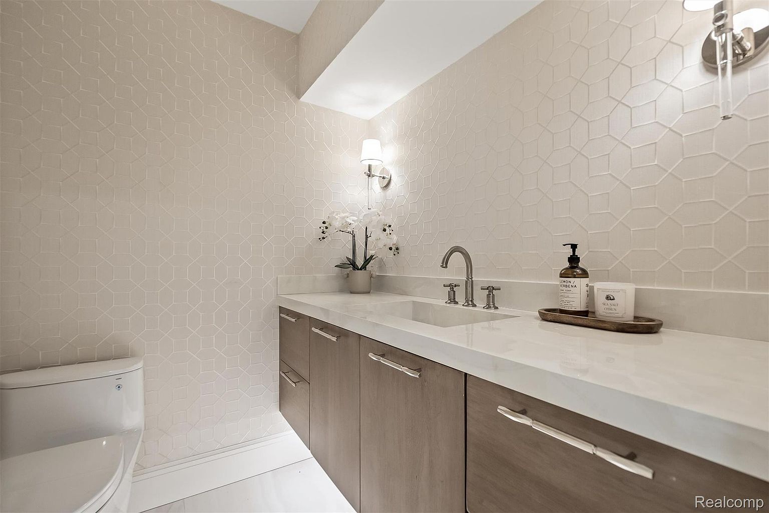 This is a well-lit guest bathroom featuring a modern design with geometric patterned wallpaper. The vanity has a white countertop and wood-grain cabinets with sleek, silver hardware. A white toilet is visible to the left, and decorative items like a soap dispenser and candle add a touch of elegance.