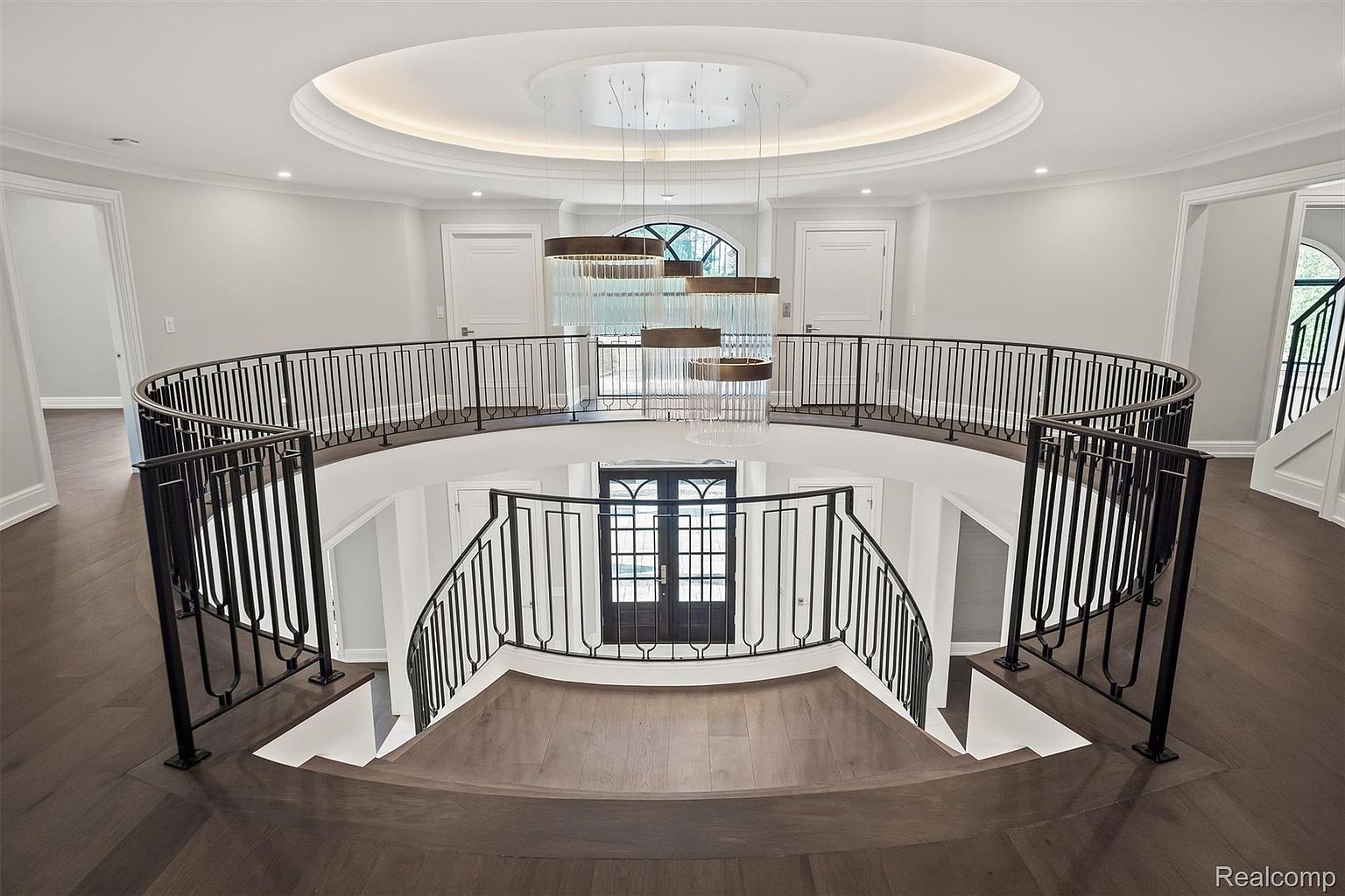 This is an interior shot of a grand foyer featuring a curved staircase and balcony overlooking the entrance. The space is illuminated by a modern chandelier hanging from a circular ceiling detail. The dark wood flooring contrasts with the light walls, creating a sophisticated and elegant atmosphere.