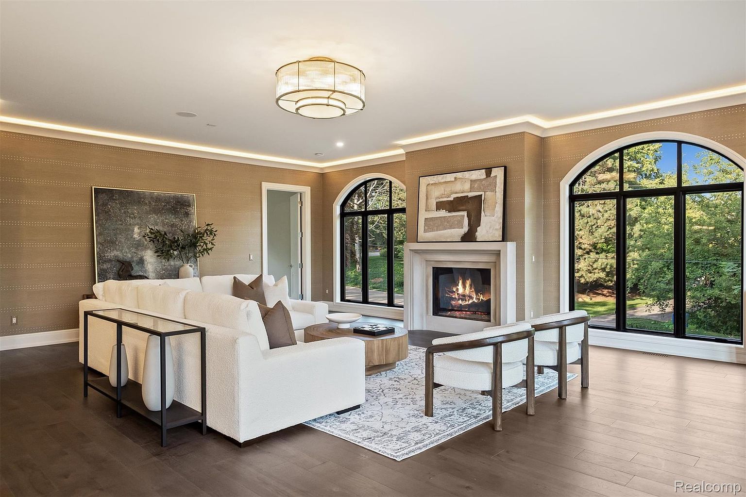This is an interior shot of a living room featuring a neutral color palette with a large white sofa, a round wooden coffee table, and two armchairs. A fireplace with an abstract painting above it serves as a focal point, flanked by arched windows that provide natural light. The room is decorated with modern accents and has hardwood flooring, creating a sophisticated and inviting atmosphere.