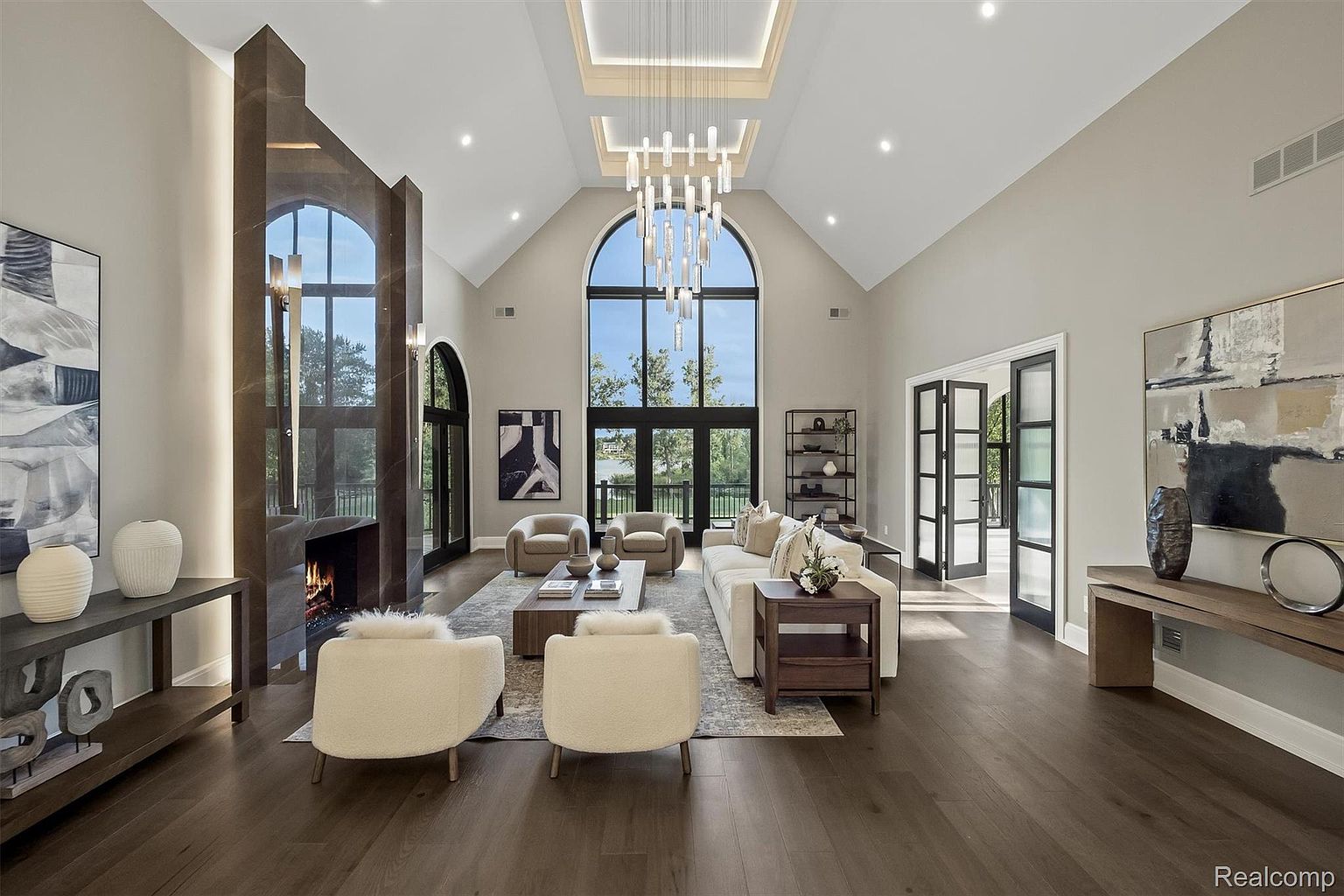 This is a spacious living room featuring high vaulted ceilings, a modern chandelier, and a large window wall that provides ample natural light. The room is furnished with a neutral-toned sofa, armchairs, and a fireplace, creating a comfortable and elegant atmosphere. Dark hardwood floors and contemporary artwork add to the room's sophisticated appeal.