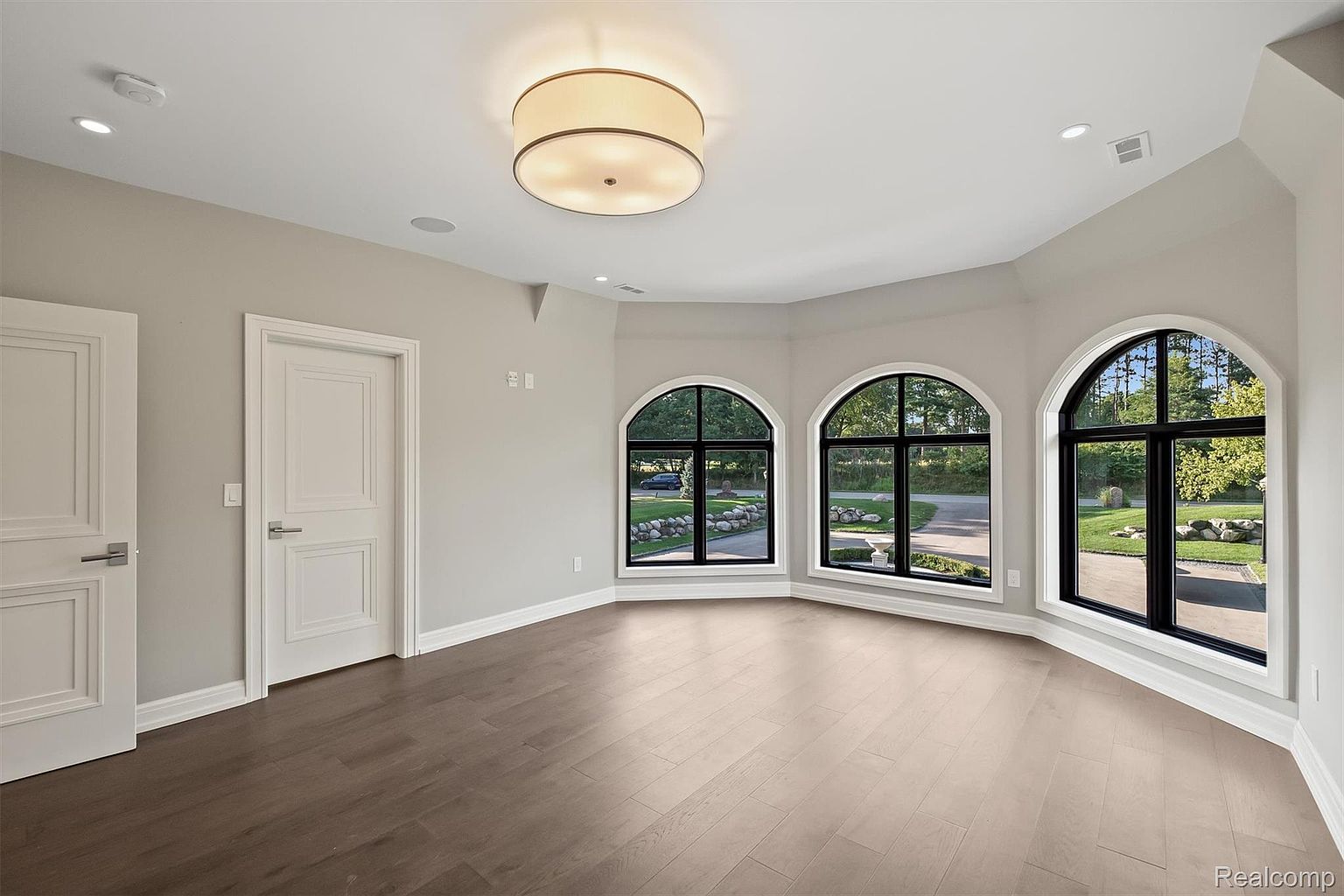 This is an interior shot of a bedroom featuring hardwood floors, light gray walls, and white trim. Three arched windows provide ample natural light and views of the exterior landscape. A modern ceiling light fixture illuminates the room, and two white paneled doors are visible on the left wall.