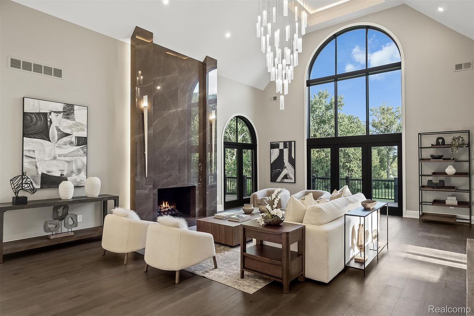 This is a luxurious living room featuring a modern fireplace with marble accents, complemented by a striking chandelier. The room is bathed in natural light from large arched windows, showcasing a view of the surrounding greenery. The neutral color palette and elegant furnishings create a sophisticated and inviting atmosphere.