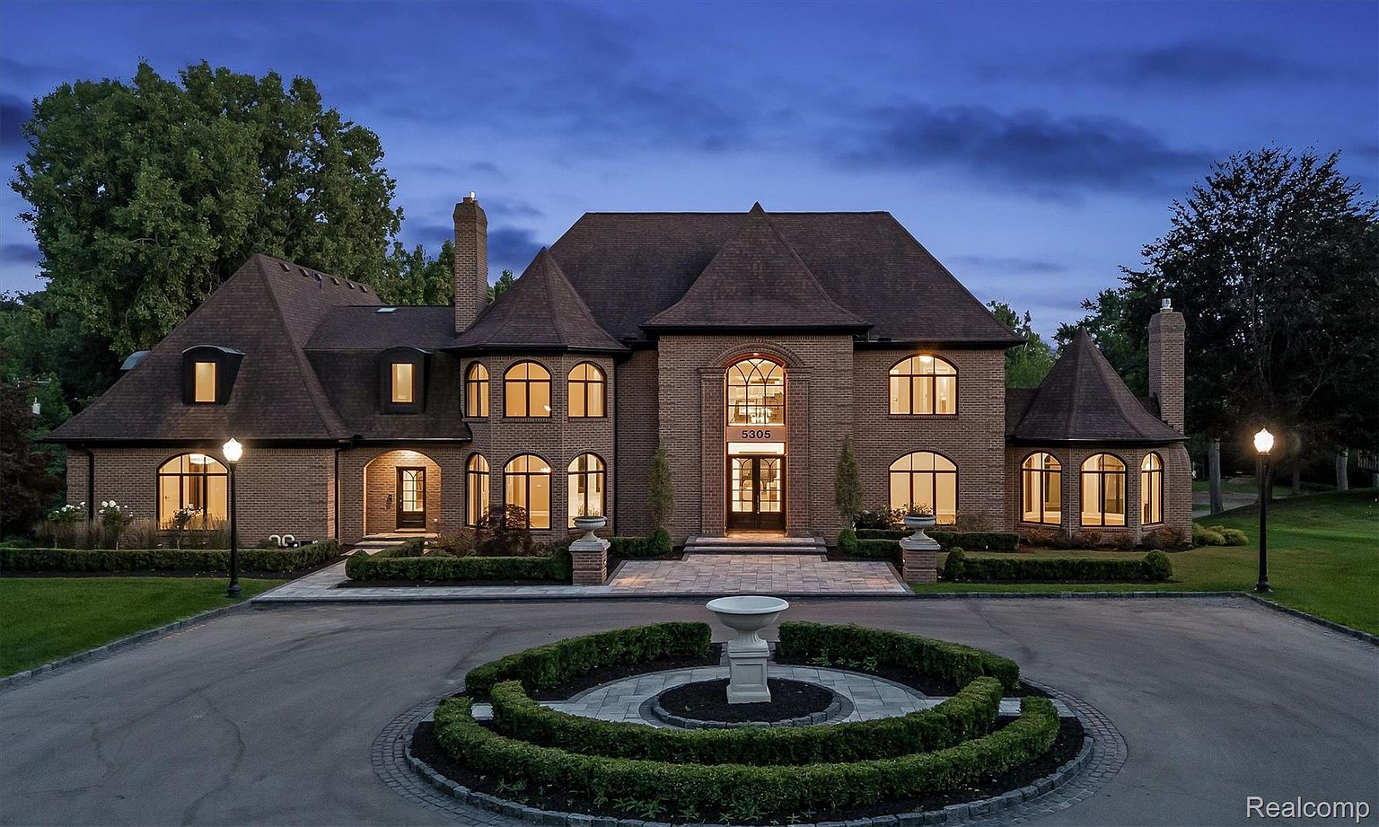 This is a front exterior view of a large, luxurious brick home at dusk. The house features a circular driveway with a fountain, manicured landscaping, and multiple arched windows that are illuminated from within. The architectural style is traditional with a grand and stately presence, suggesting high-end real estate.