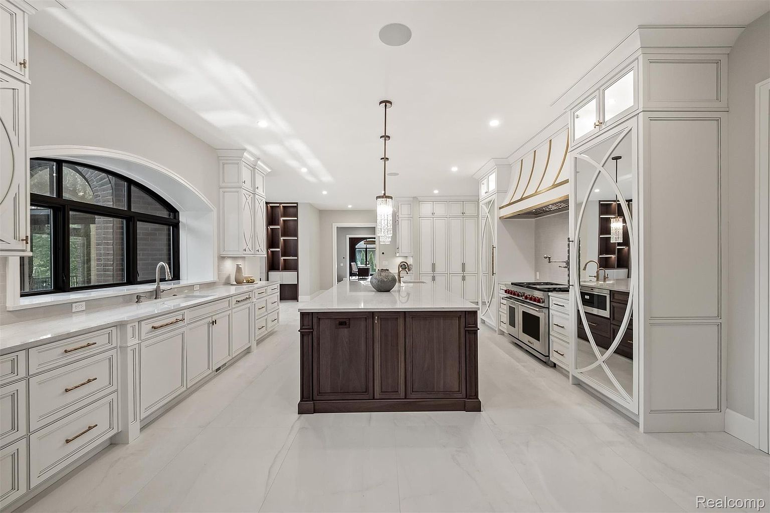 This is a spacious, high-end kitchen featuring custom white cabinetry with detailed trim, a dark wood island with a marble countertop, and high-end stainless steel appliances. The kitchen is well-lit with recessed lighting and a decorative pendant light above the island, and the overall design is elegant and luxurious. The perspective is a wide shot, showcasing the entire kitchen layout and its high-end finishes.