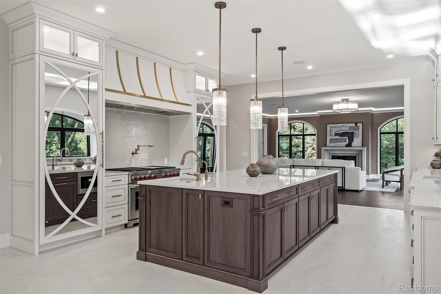 This is a beautifully designed kitchen featuring a large center island with dark wood cabinetry and a white countertop. The kitchen also includes custom white cabinetry with mirrored accents, high-end appliances, and elegant pendant lighting. The open layout connects to a living area, creating a seamless flow and a luxurious feel.