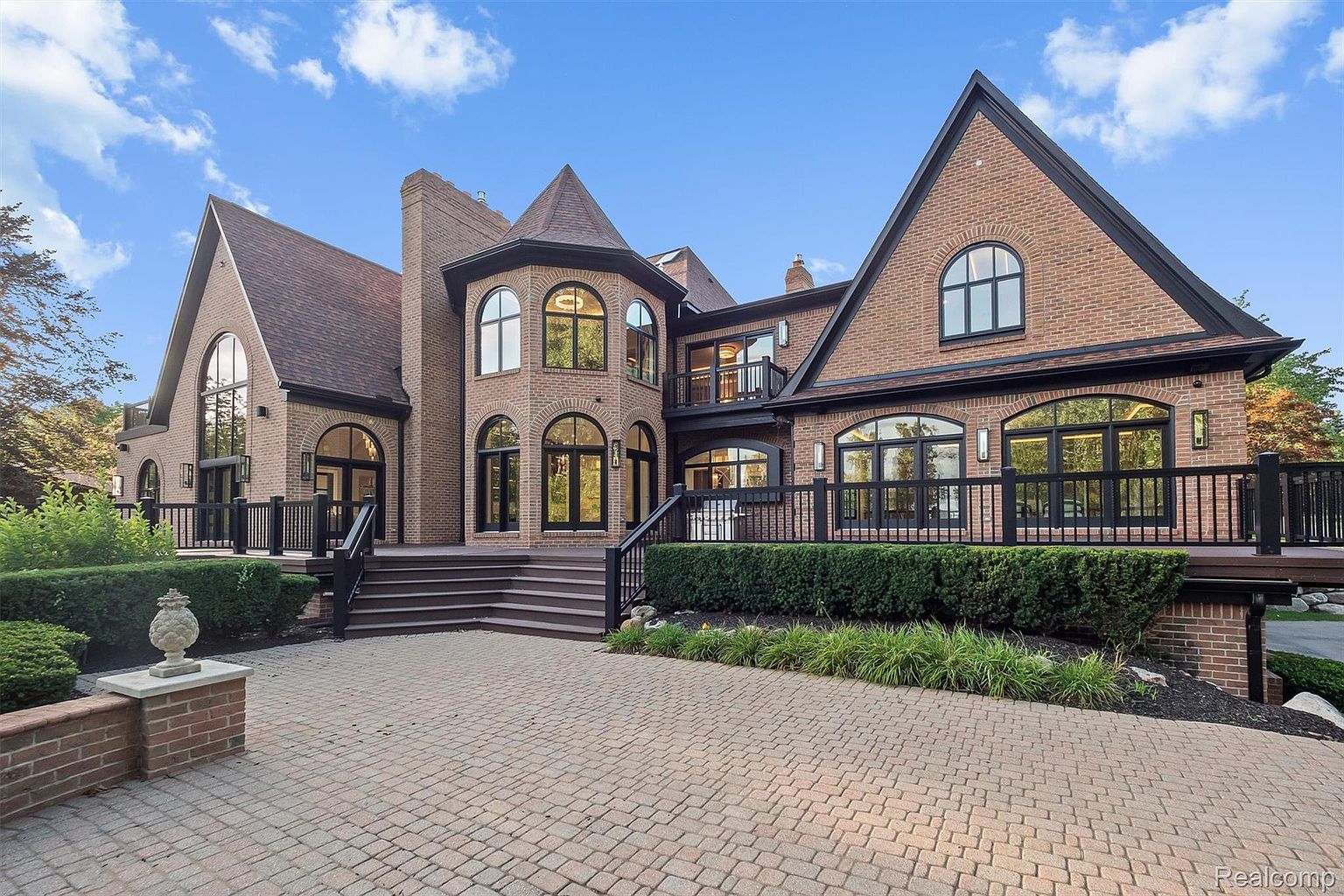 This is a front exterior view of a large, luxurious brick home. The house features multiple gabled roofs, arched windows with black frames, and a spacious brick driveway. A wooden deck with black railings extends across part of the front, and manicured landscaping adds to the property's curb appeal.